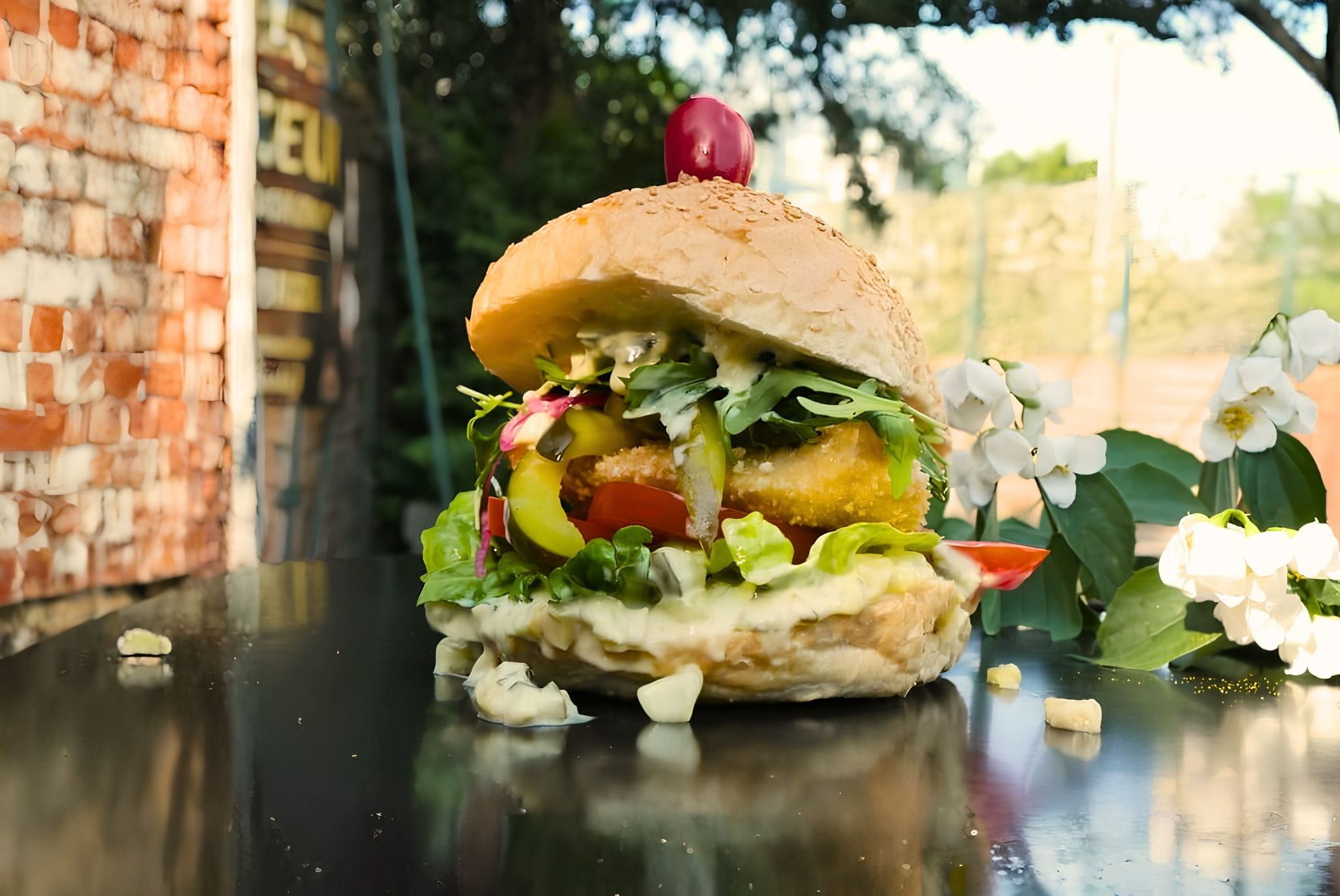 Duży burger z warzywami, piklami i sosem na czarnym tle z kwiatami w tle.