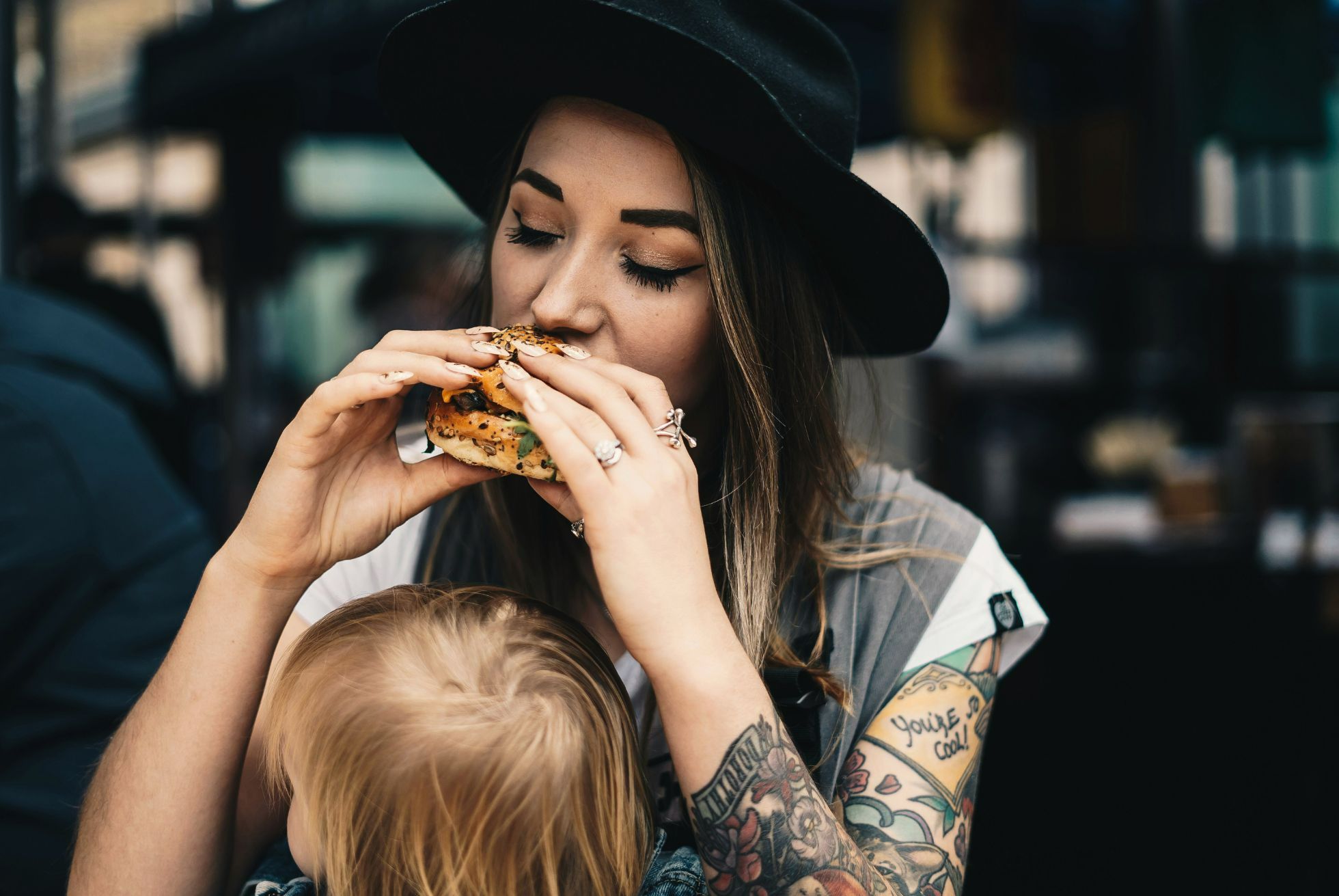 Kobieta w kapeluszu trzyma burgera, podczas gdy małe dziecko siedzi na jej kolanach.