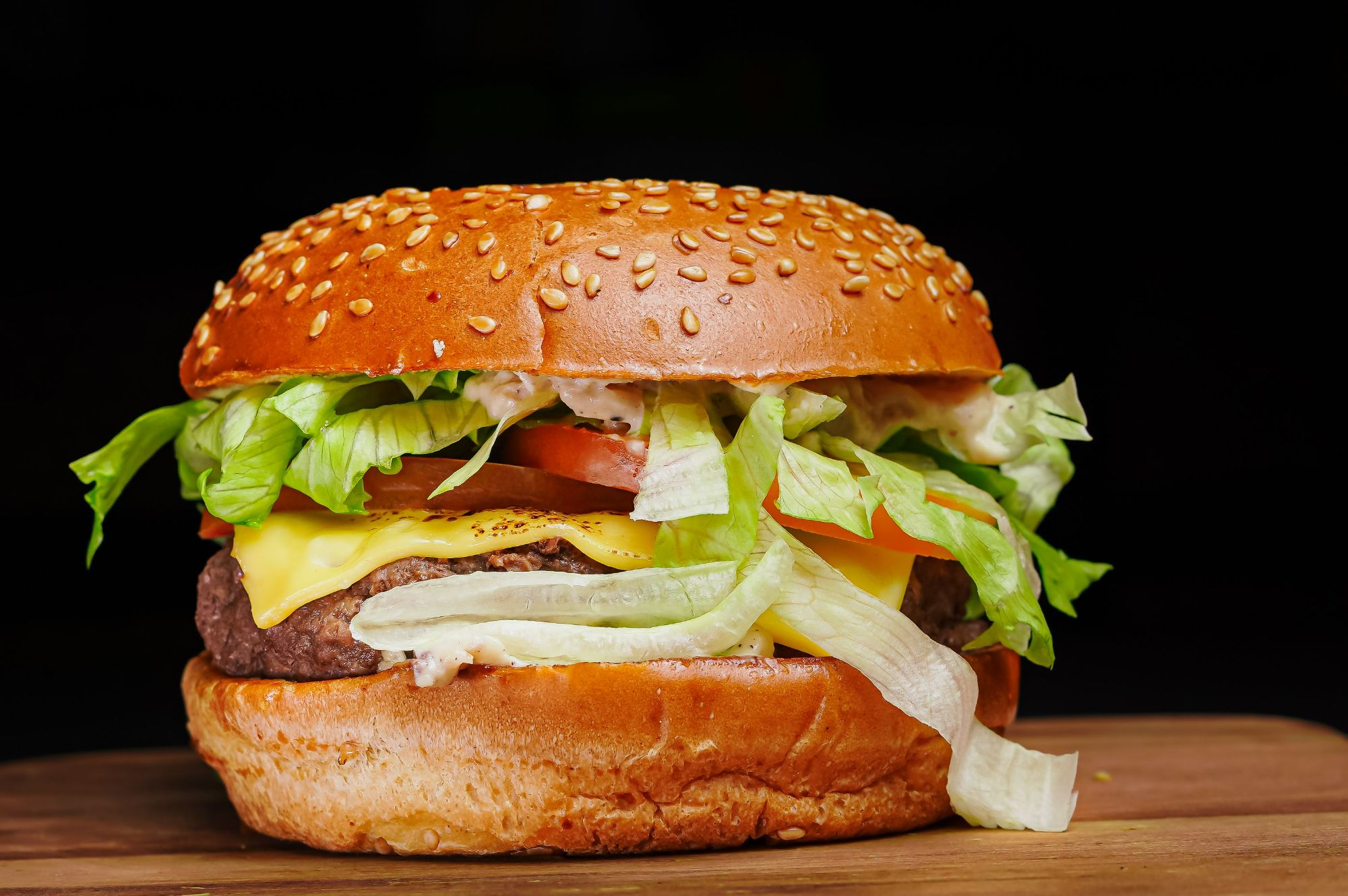 Soczysty hamburger z serem, sałatą, pomidorem i sezamowym pieczywem na drewnianym tle.