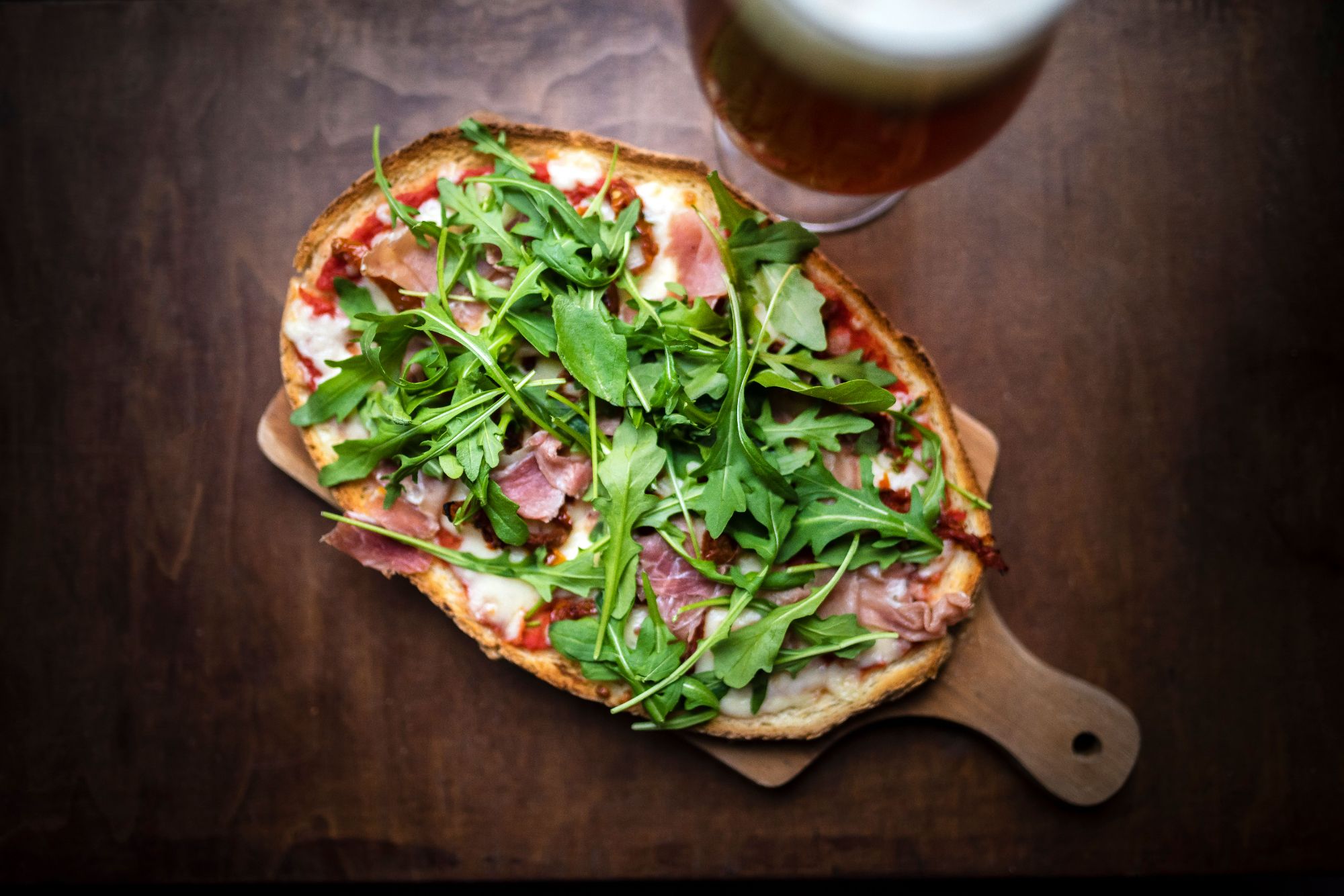 Pizza z szynką, serem i rukolą, podana na desce z piwem w tle.