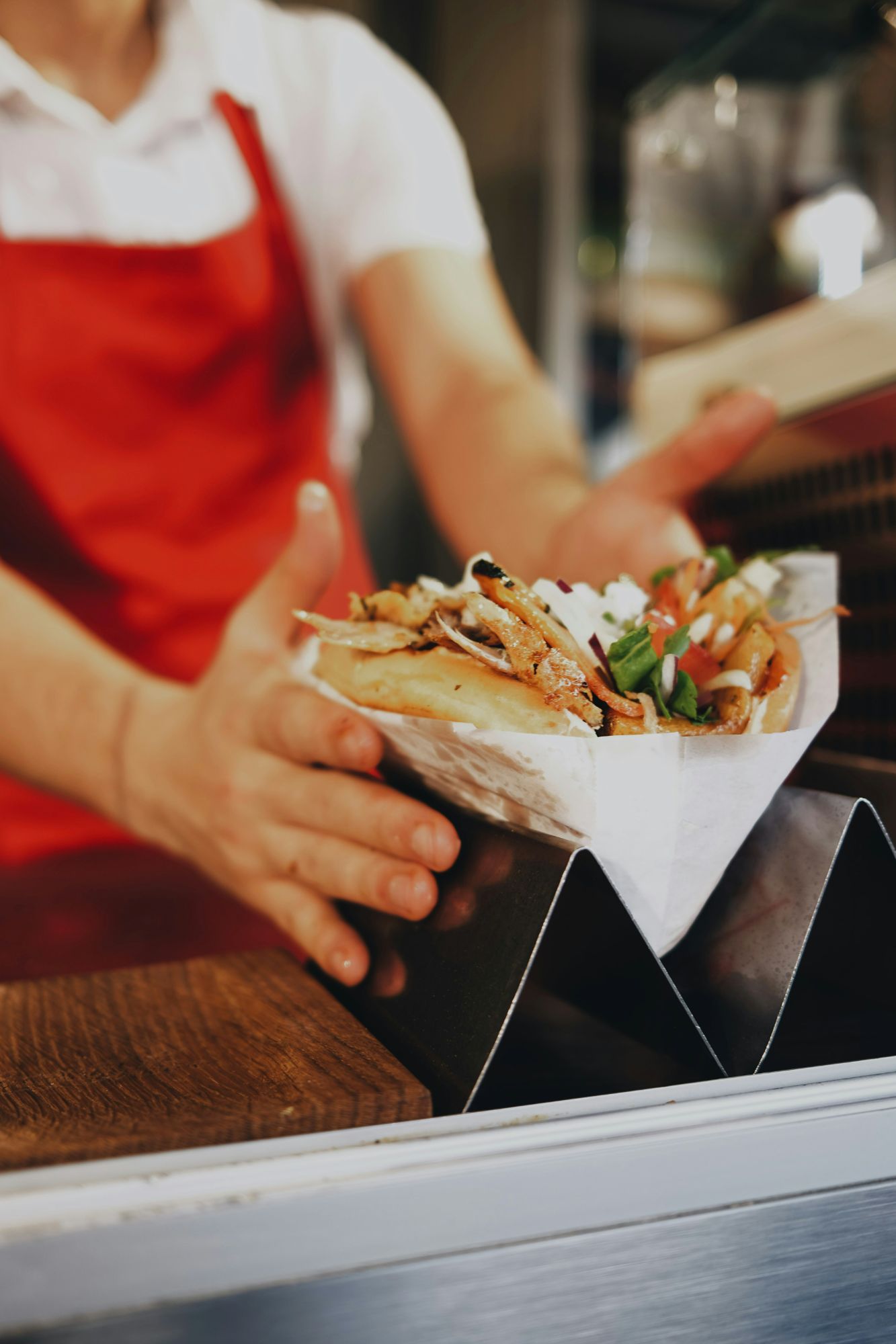 Sprzedawca serwuje kebab w papierowym opakowaniu, trzymając je w rękach, z dodatkami takimi jak sałata i pomidory.