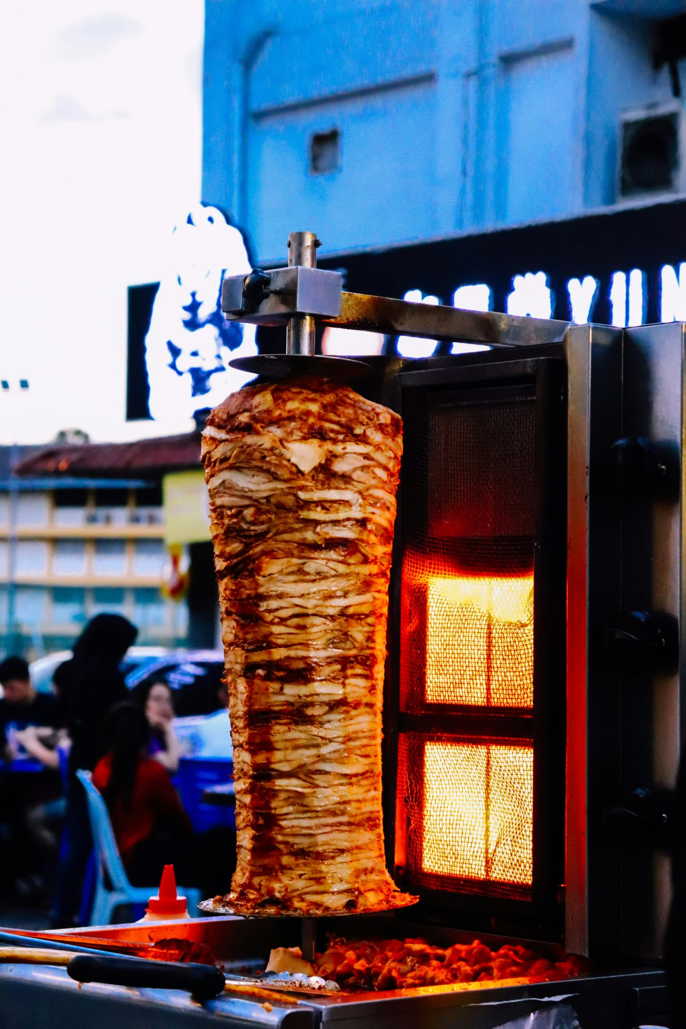 Kebab na rożnie, grillowany z kolorowymi sosami w tle, przy ulicznym stoisku z jedzeniem.