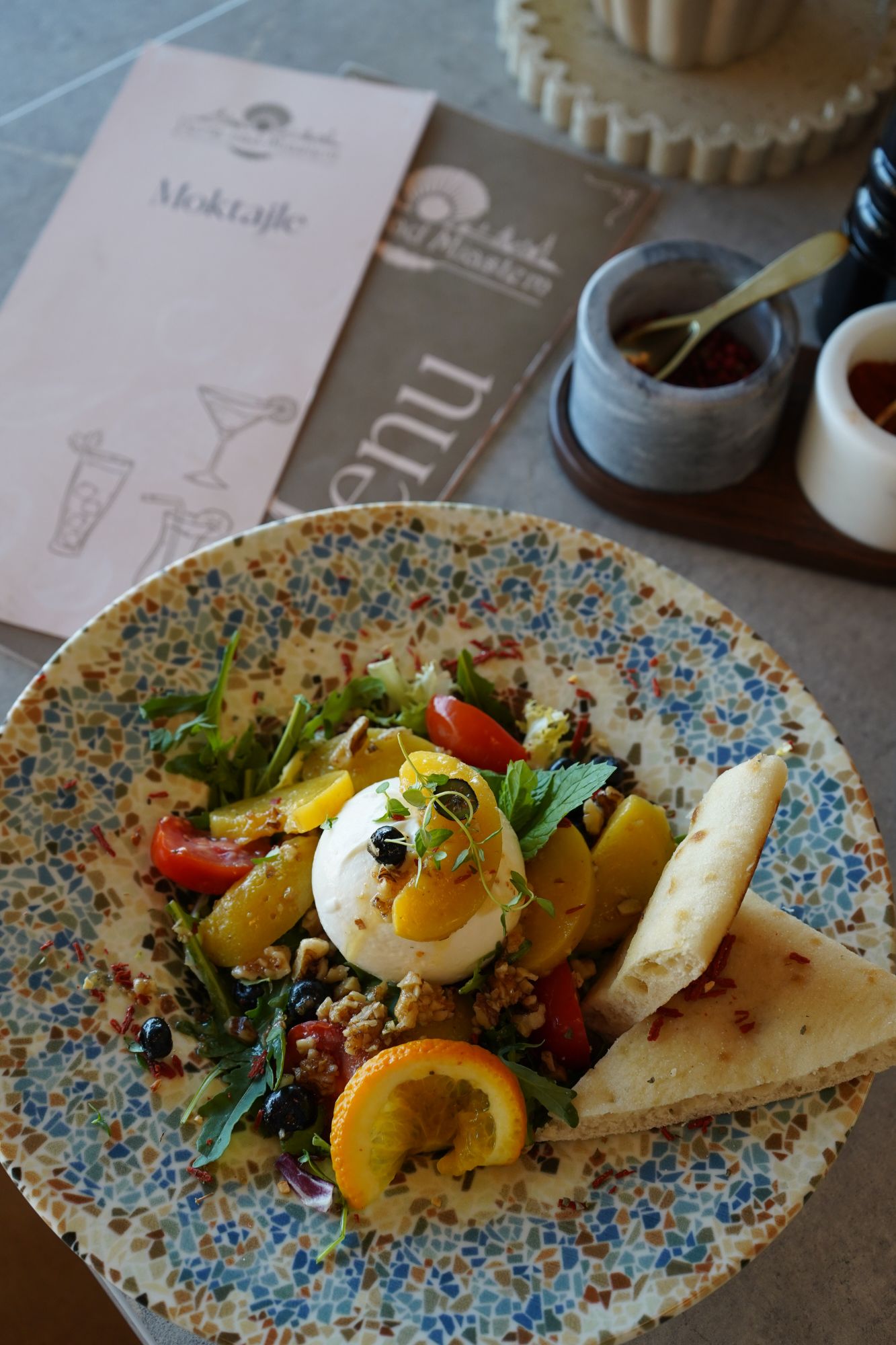 Sałatka z serem burrata, warzywami i owocami na talerzu, w tle menu kawiarni i akcesoria do serwowania.