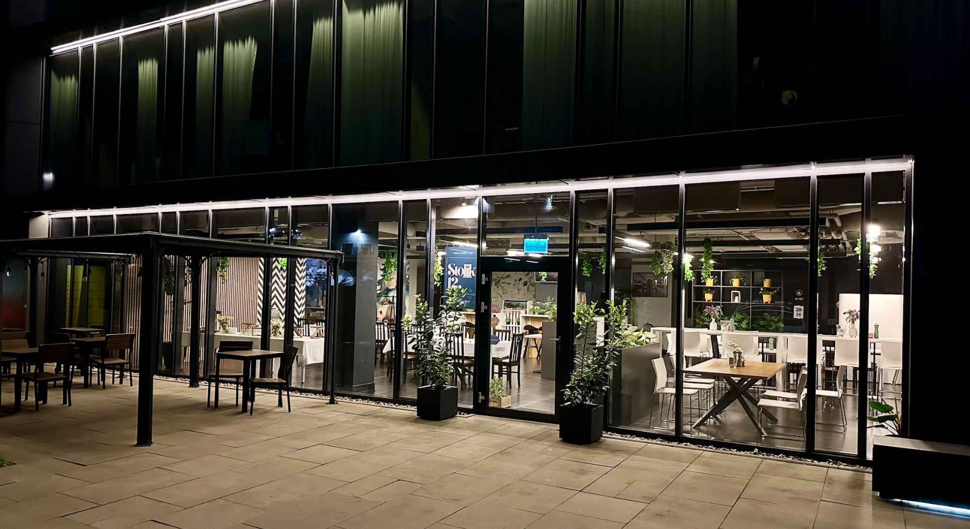 Widok na nowoczesną restaurację nocą, z dużymi przeszklonymi witrynami i otwartą przestrzenią.