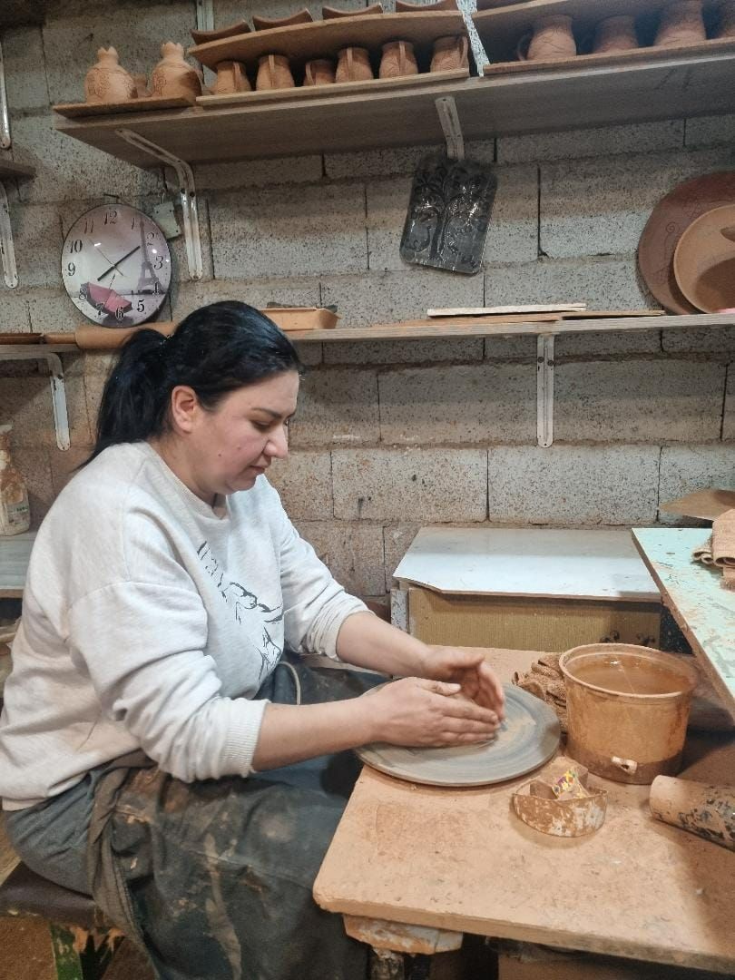 Kobieta lepiąca ceramikę na kole garncarskim w warsztacie, z półkami pełnymi naczyń w tle.