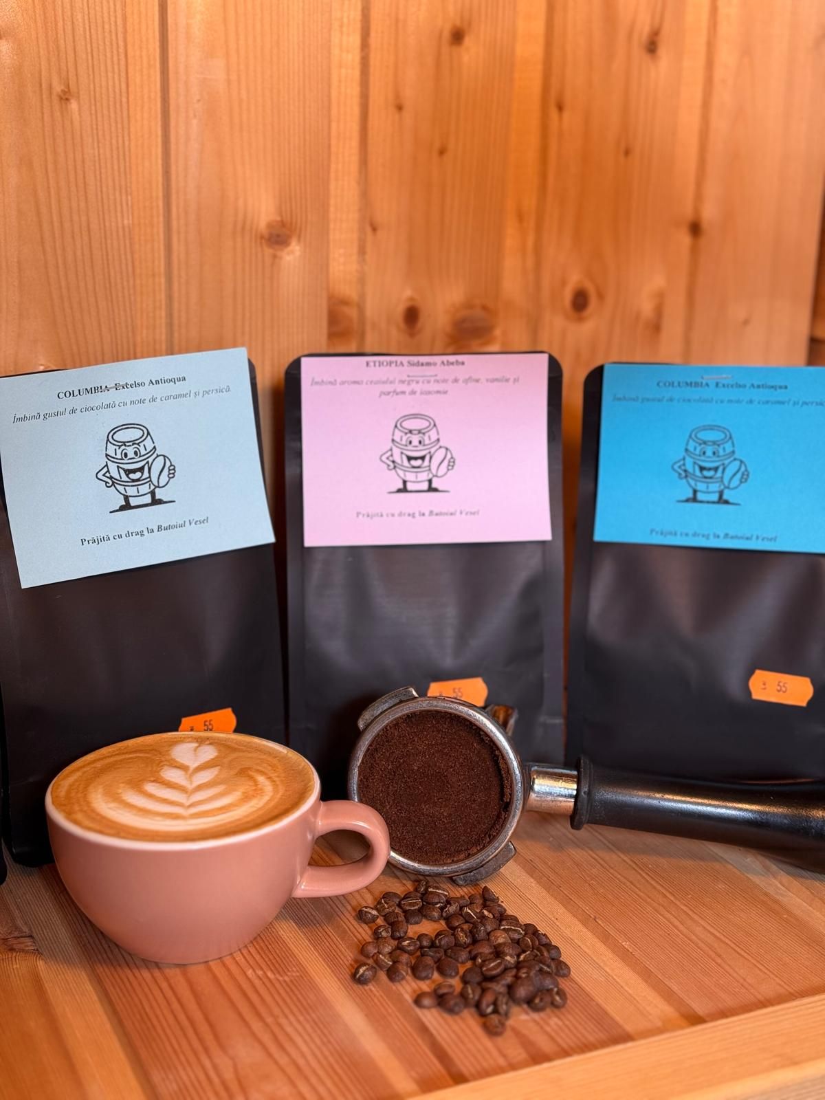 Cafetieră cu trei pungi de cafea, o ceșcă de cafea cu lapte și boabe de cafea pe o masă din lemn.