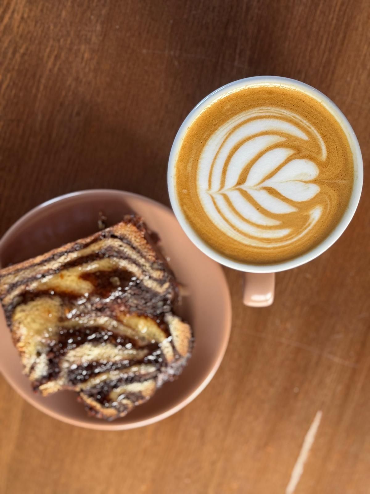 O ceașcă de cafea cu model de inimă alături de o felie de cozonac zebra pe un background de lemn.