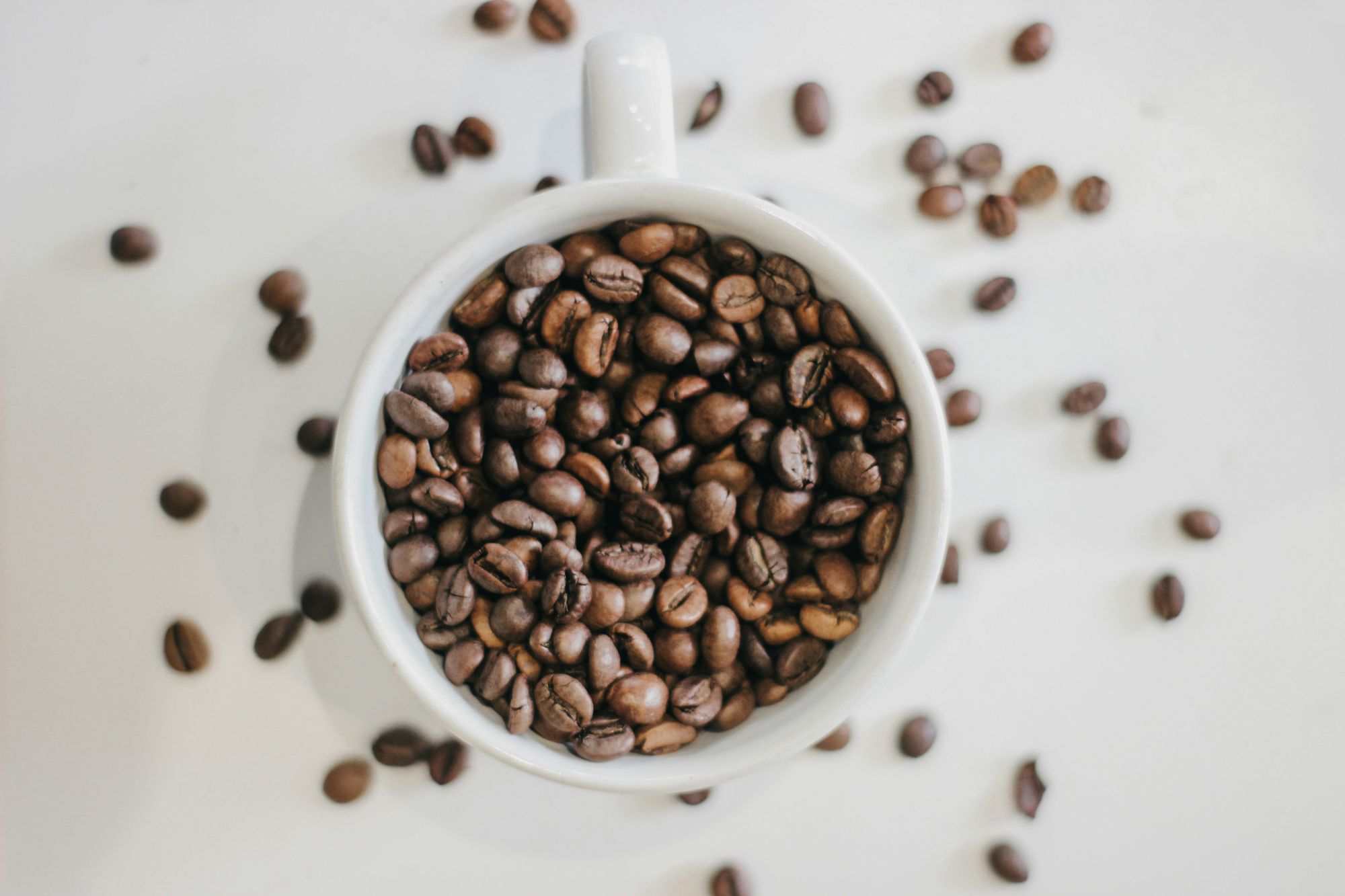 Cupa albă plină cu boabe de cafea prăjite, înconjurată de boabe dispersate pe o suprafață albă.