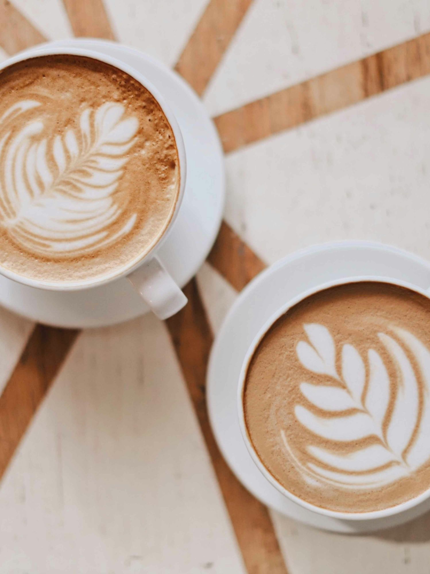 Două căni de cafea cu latte art, plasate pe o masă cu un model geometric din lemn.