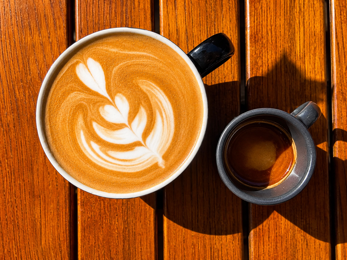 Cafetieră mare cu latte art și un espresso mic, pe o masă din lemn.