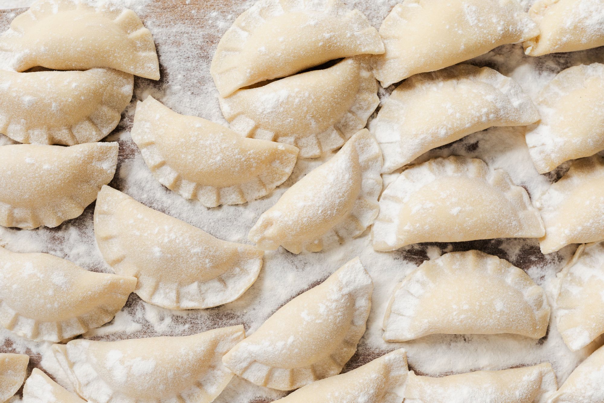 Surowe pierogi na blasze, posypane mąką, w różnych kształtach i rozmiarach.