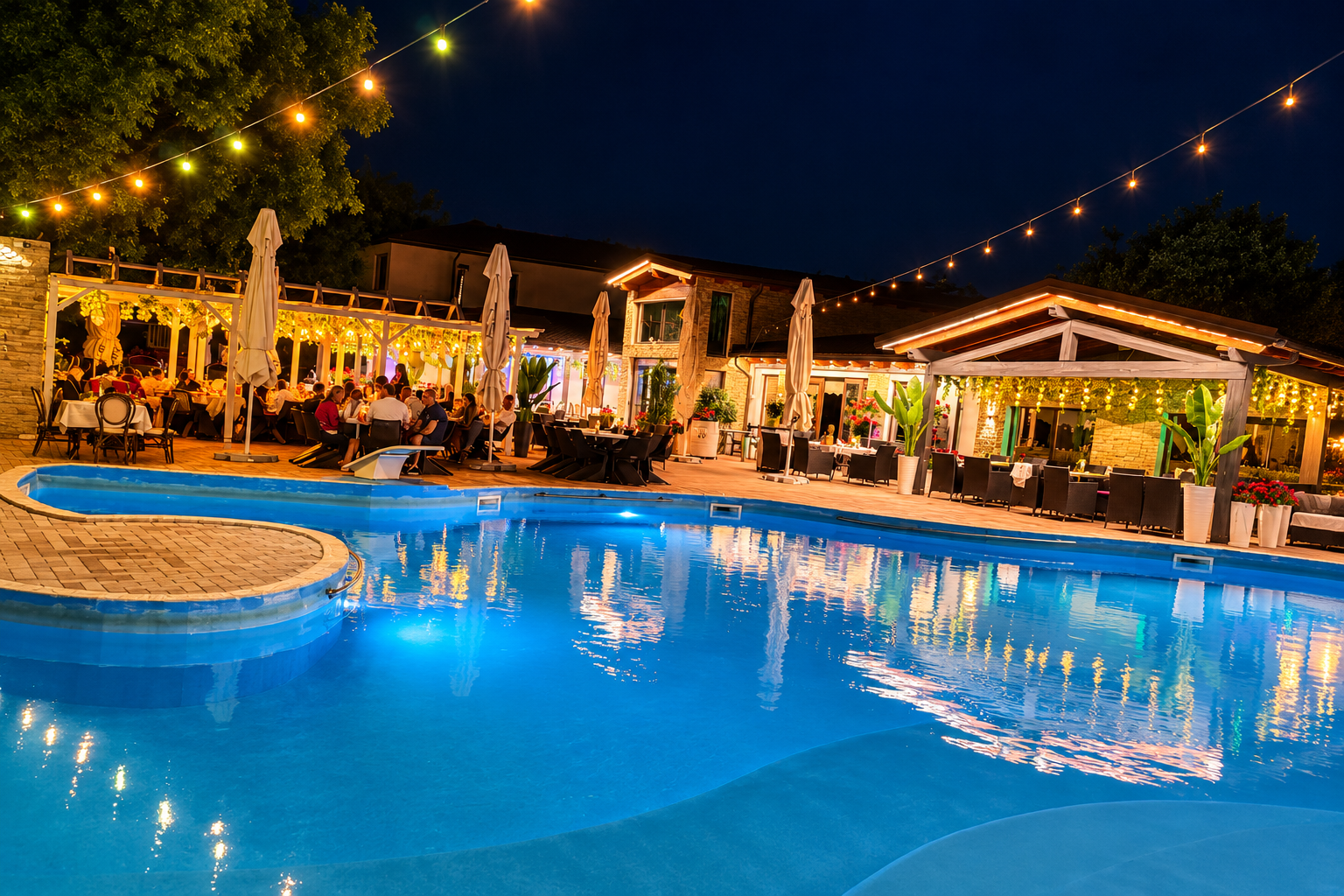Piscină iluminată noaptea, cu mese și umbrele, oameni socializând, decor festiv cu lumini.