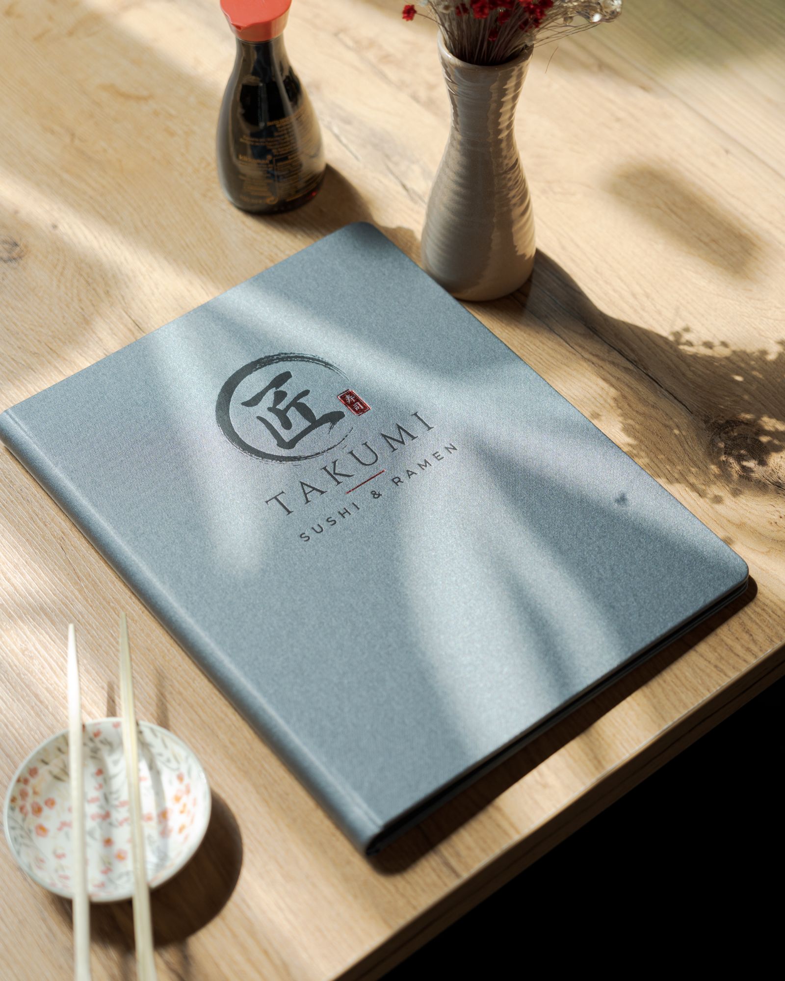Menu restauracji Takumi, sushi i ramen, leżące na drewnianym stole obok pałeczek i sosu sojowego.