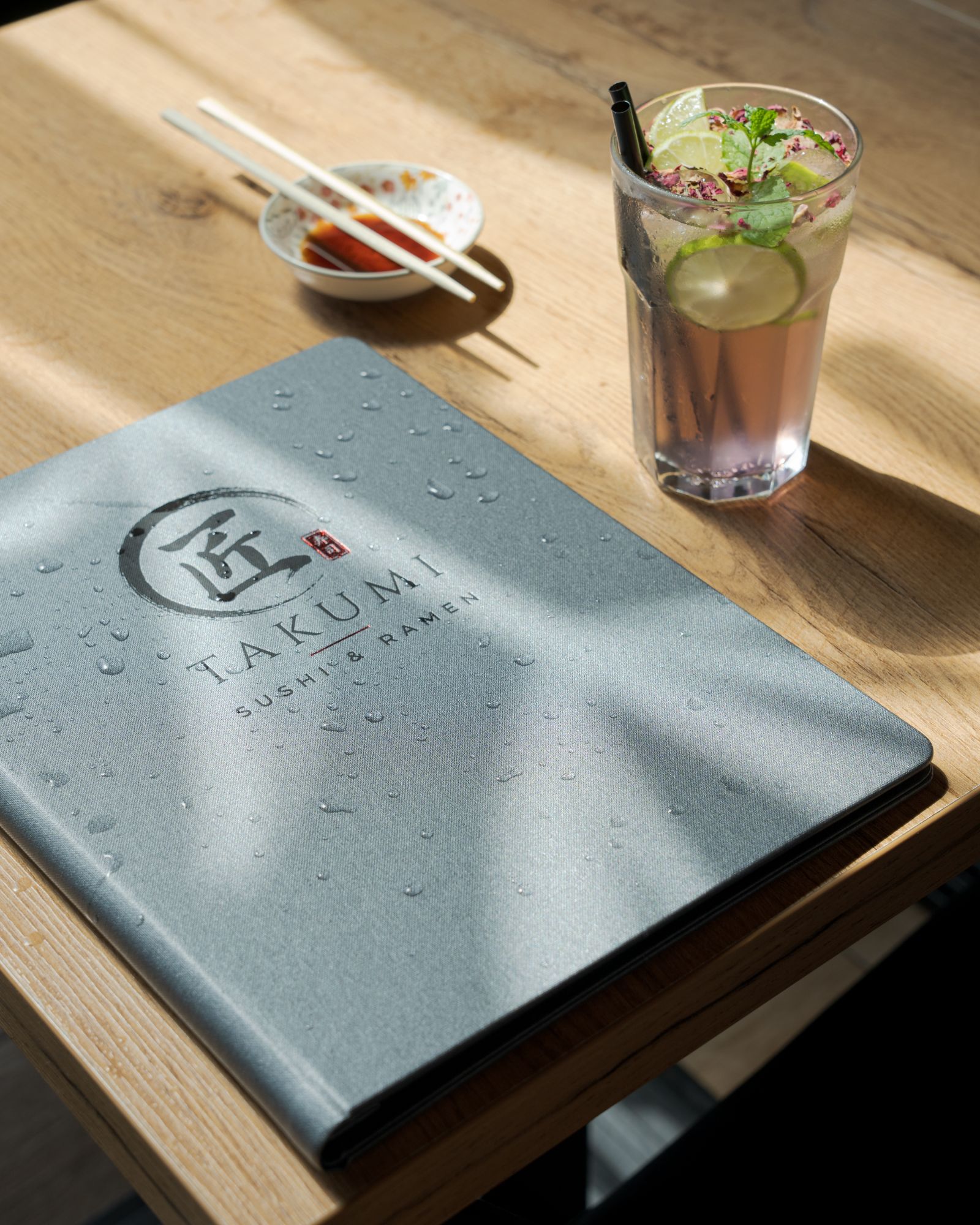 Menu restauracji Takumi, sushi i ramen, obok szklanka z napojem i talerz z sosem na drewnianym stole.