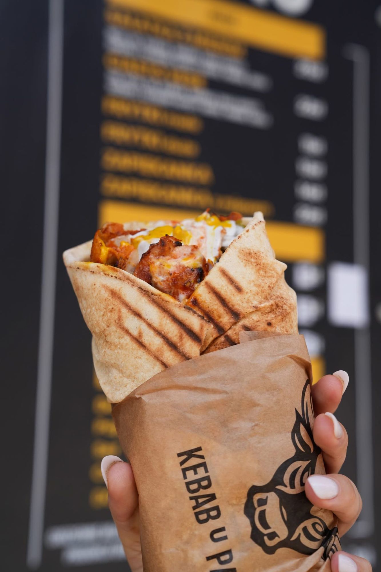 Kebab w wrapie trzymany w ręku, w tle menu z potrawami.