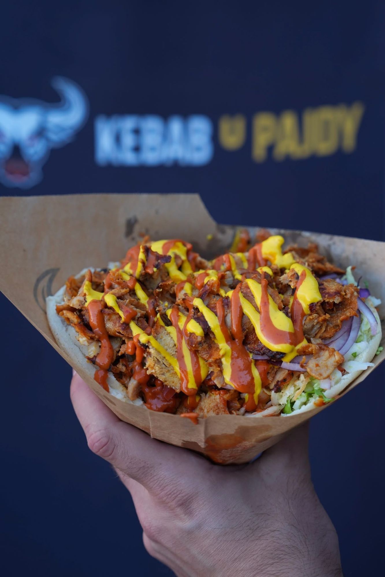 Ręka trzymająca kebab z mięsem, surówką i sosami, na tle logo Kebab u Pajdy.