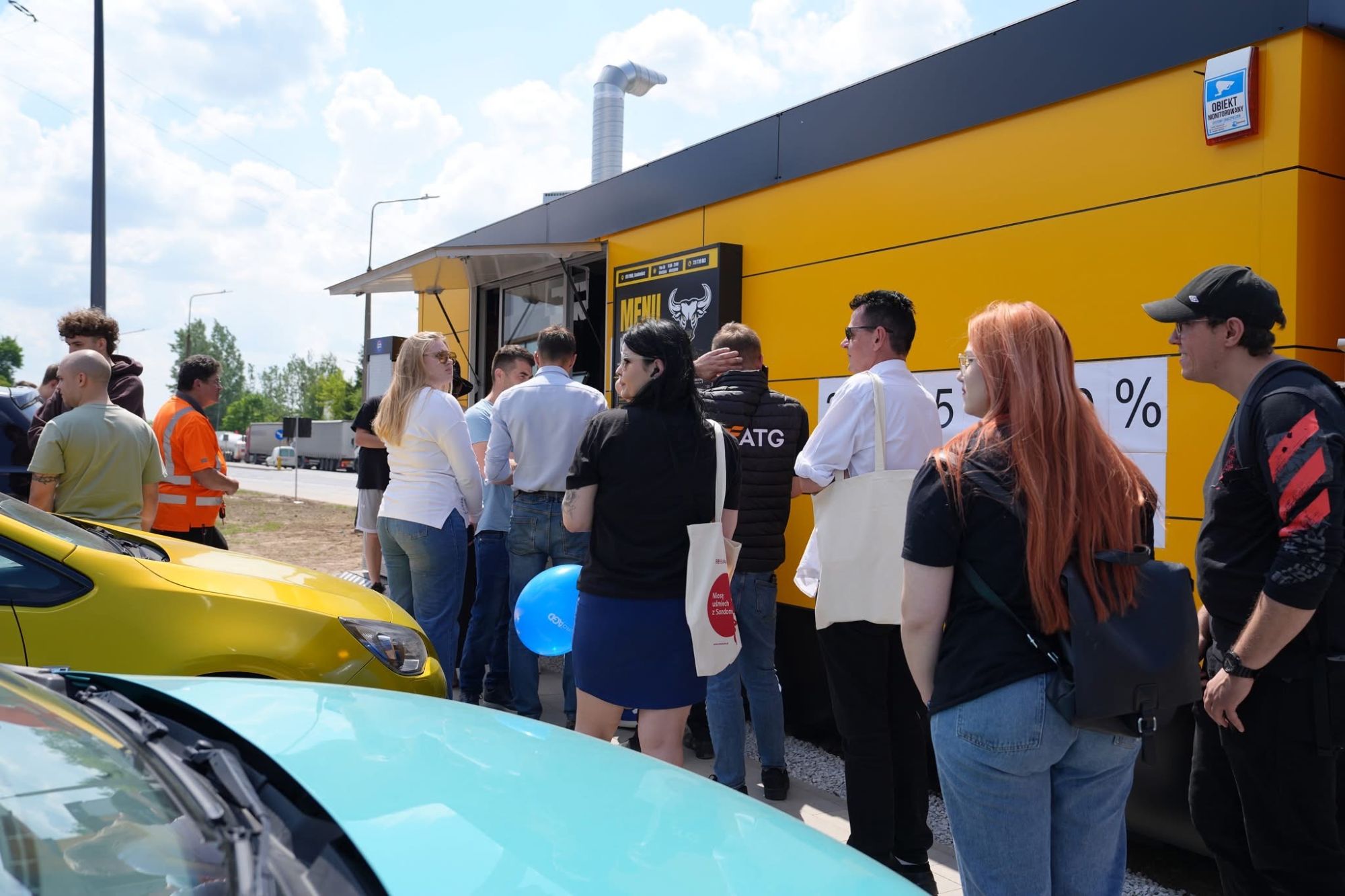 Ludzie stoją w kolejce przed żółtym punktem gastronomicznym, czekając na jedzenie lub napój.