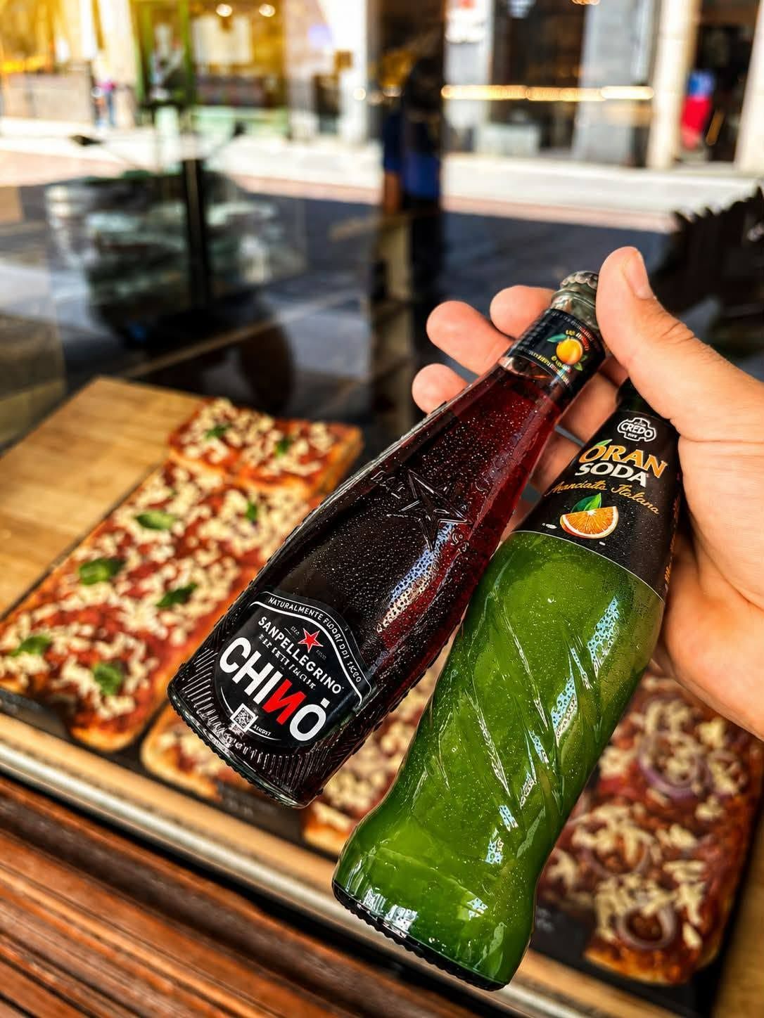 Butelki napojów: ciemna butelka Chino oraz jasnozielona Gran Sorega, w tle pizza w witrynie sklepu.
