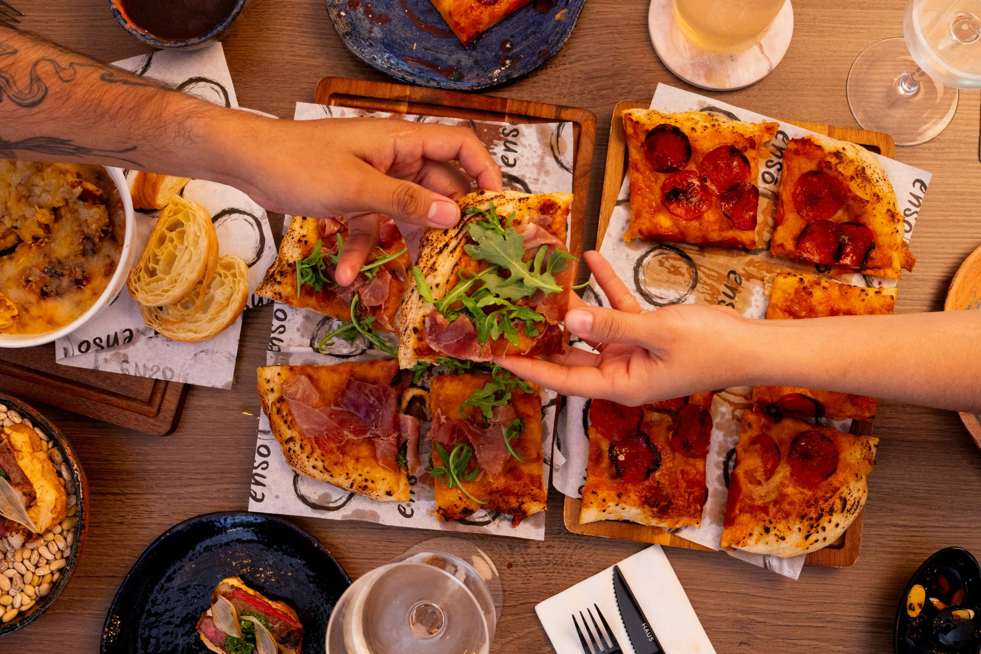Ręce sięgają po pizzę na drewnianych deskach, wokół jedzenie i napoje na stole.