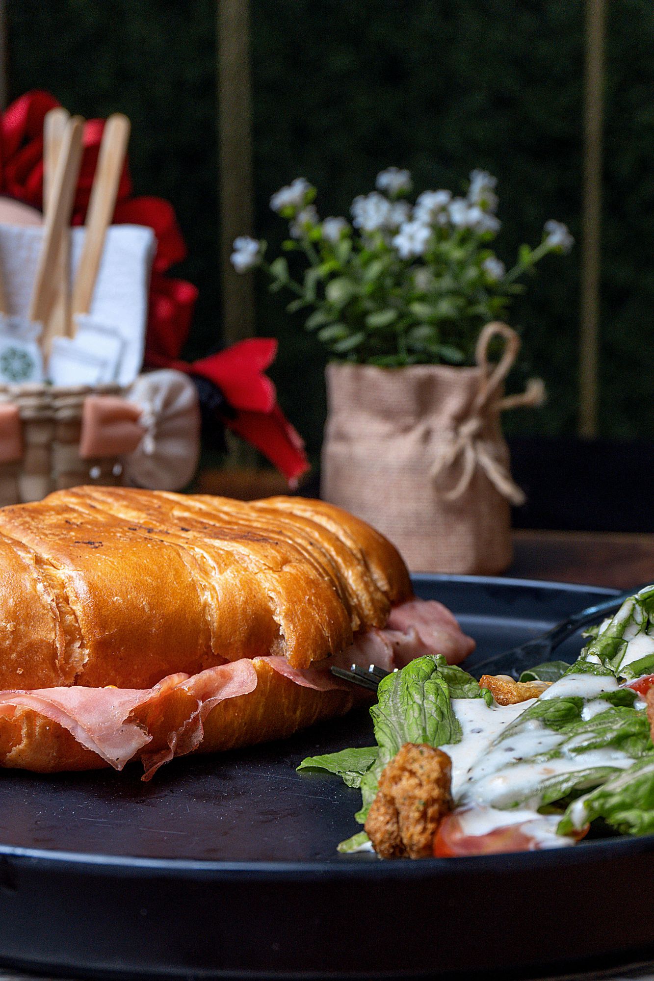 Złocisty croissant z szynką podany z sałatką warzywną i sosem, w tle dekoracje i kwiaty.