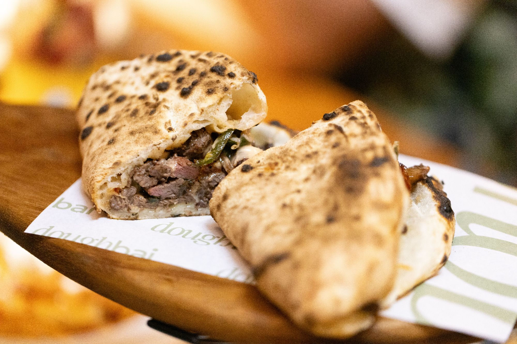 Smażona tortilla z mięsem, sera i warzywami, trzymana na drewnianej desce, w tle nieostre jedzenie.