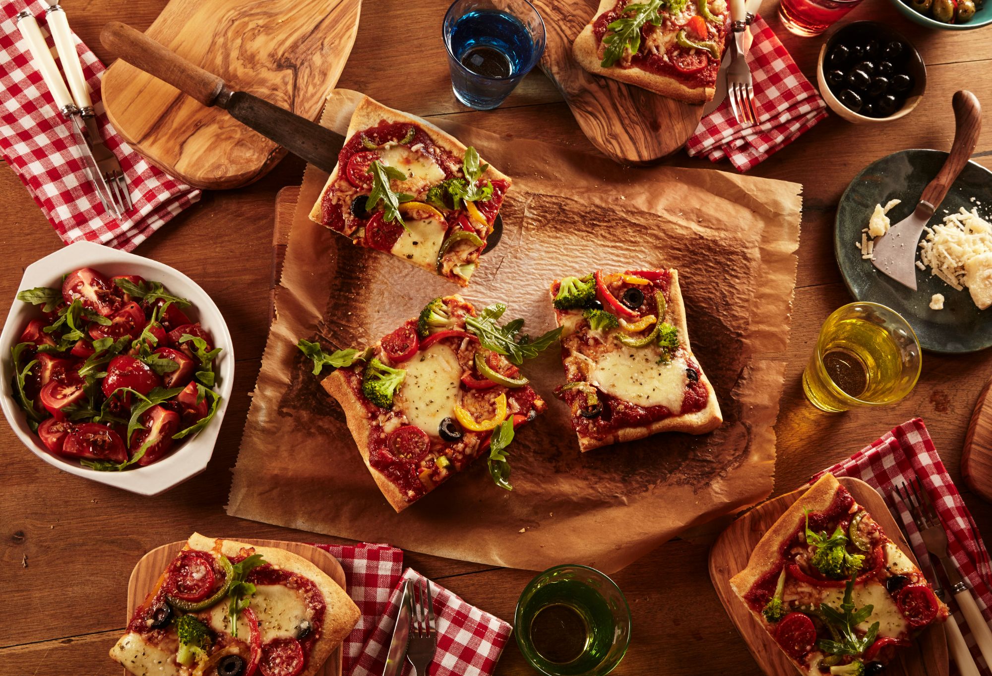 Pizza z serem i warzywami na drewnianym stole, w otoczeniu sałatki oraz kolorowych szklanek.