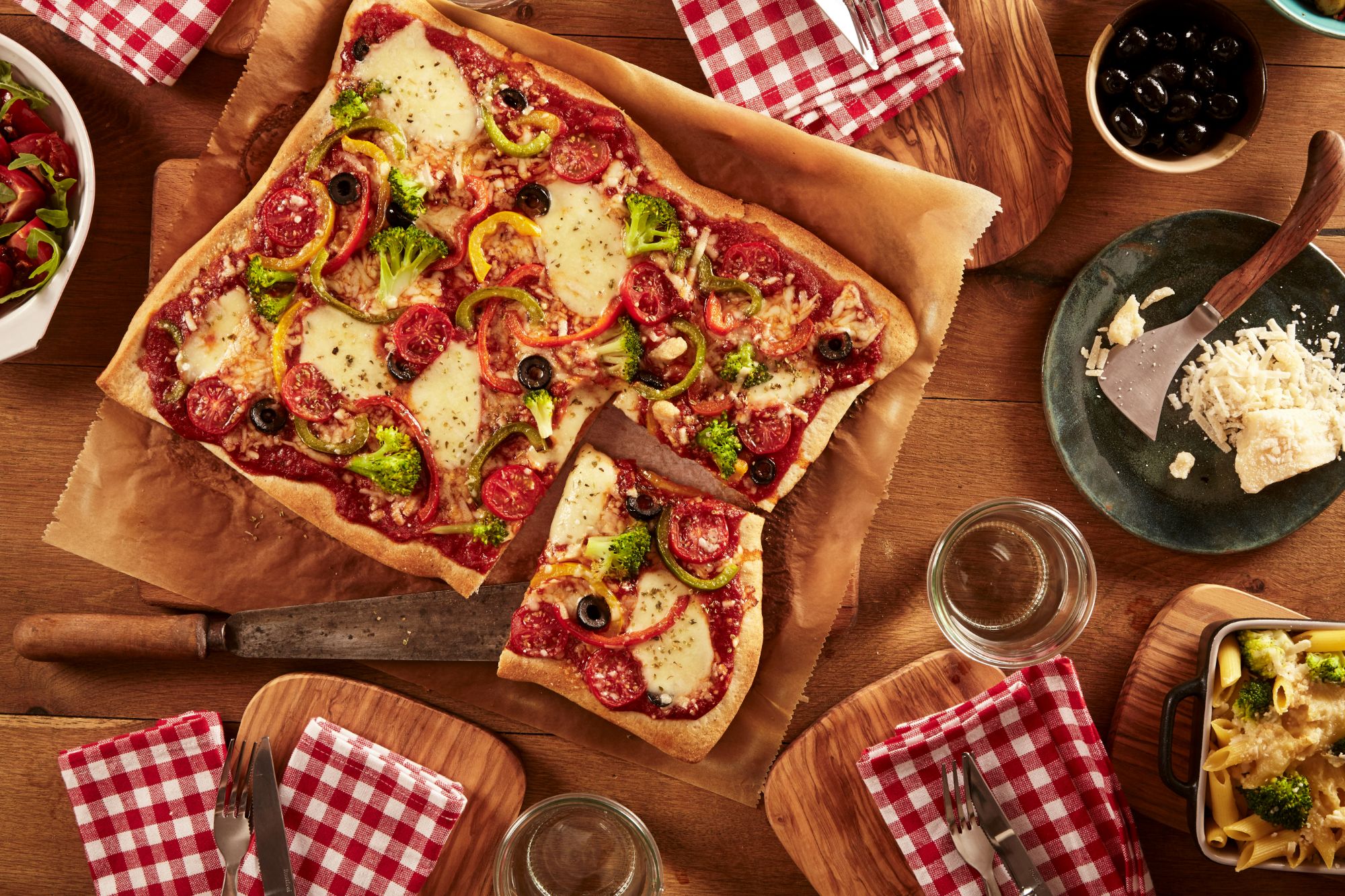 Pizza z serem, pepperoni, warzywami, podana na drewnianym blacie z dodatkowymi składnikami i zastawą stołową.