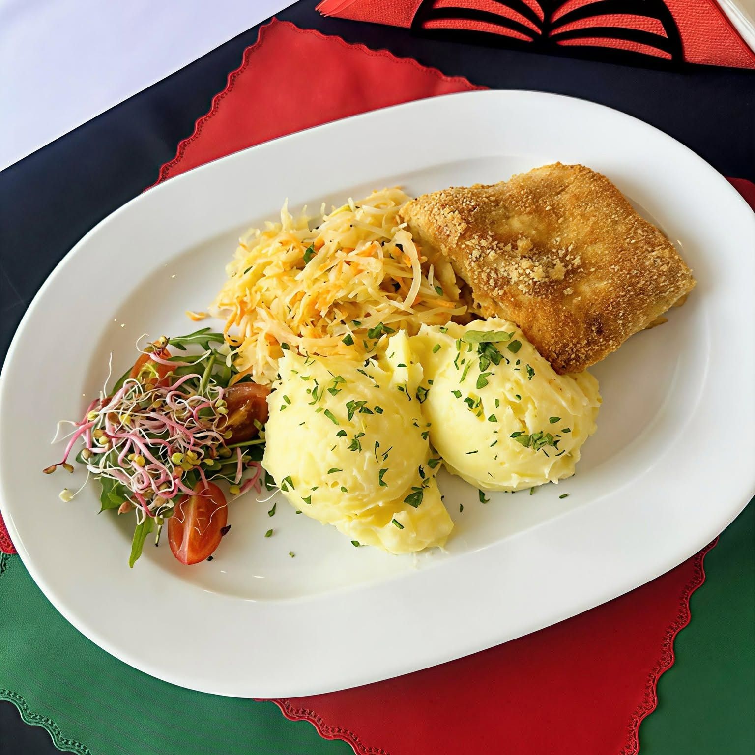 Talerz z puree ziemniaczanym, rybą, surówką i kiełkami, udekorowany ziołami na kolorowym obrusie.