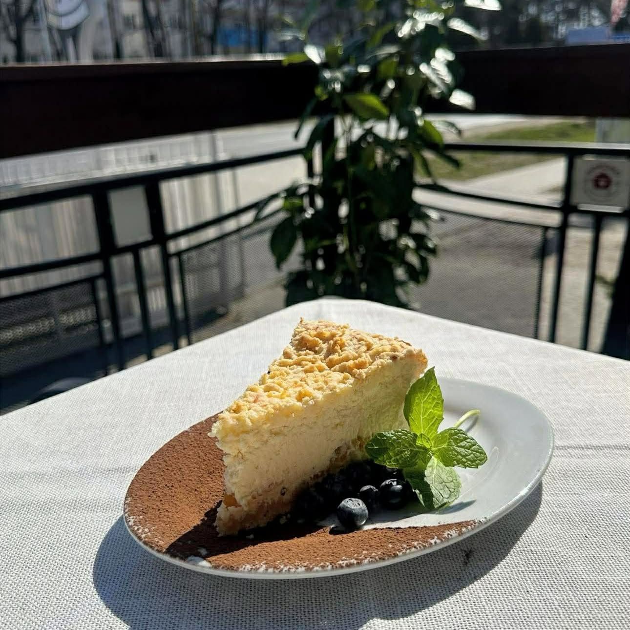 Kawałek sernika na talerzu z miętą i jagodami, na tle zewnętrznej kawiarni w słoneczny dzień.