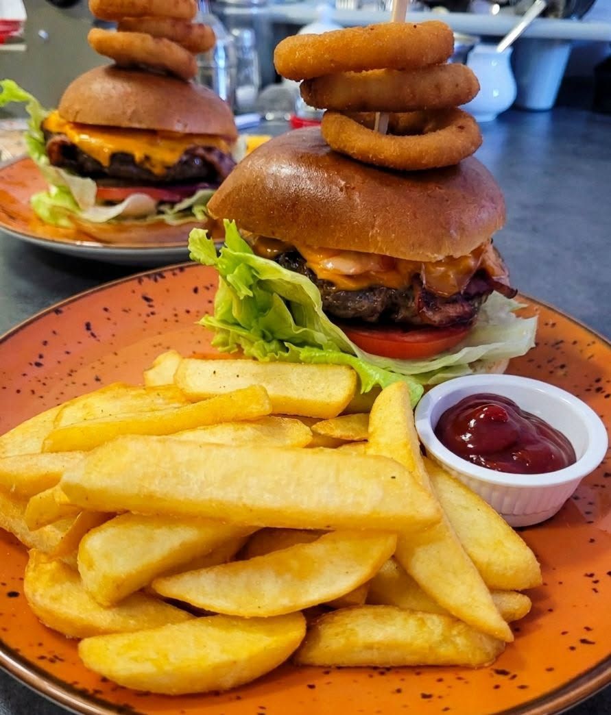 Burger z serem, sałatą i pomidorem, z krążkami cebuli na wierzchu, podany z frytkami i ketchupem.