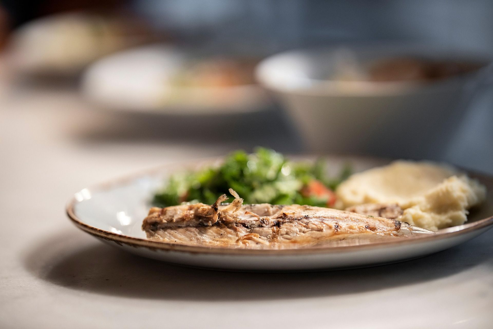 Talerz z grillowaną rybą, sałatką i puree ziemniaczanym, z rozmytymi daniami w tle.