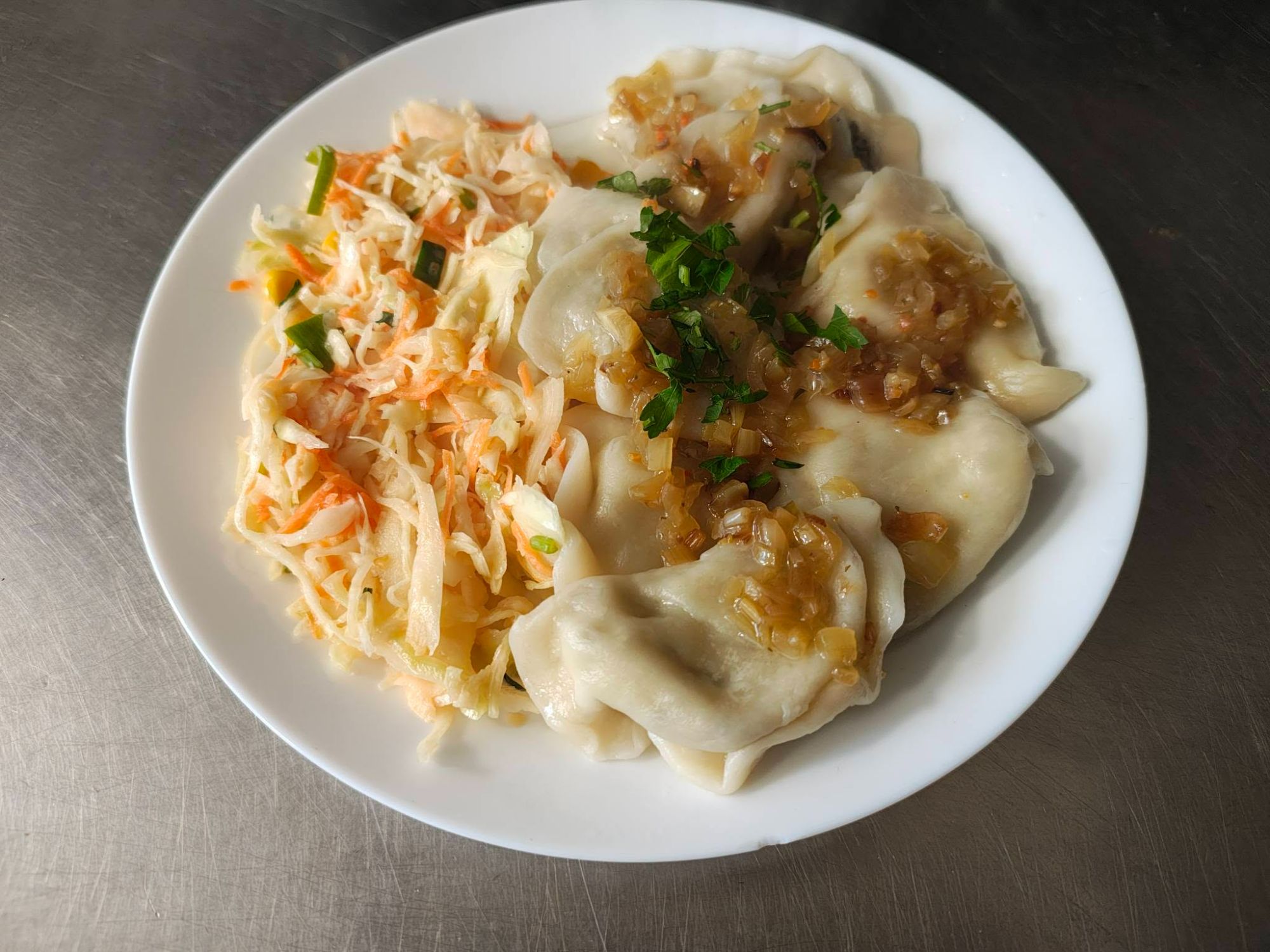 Pierogi na talerzu z sosem, posypane ziołami, obok surówka z kapusty i marchewki.