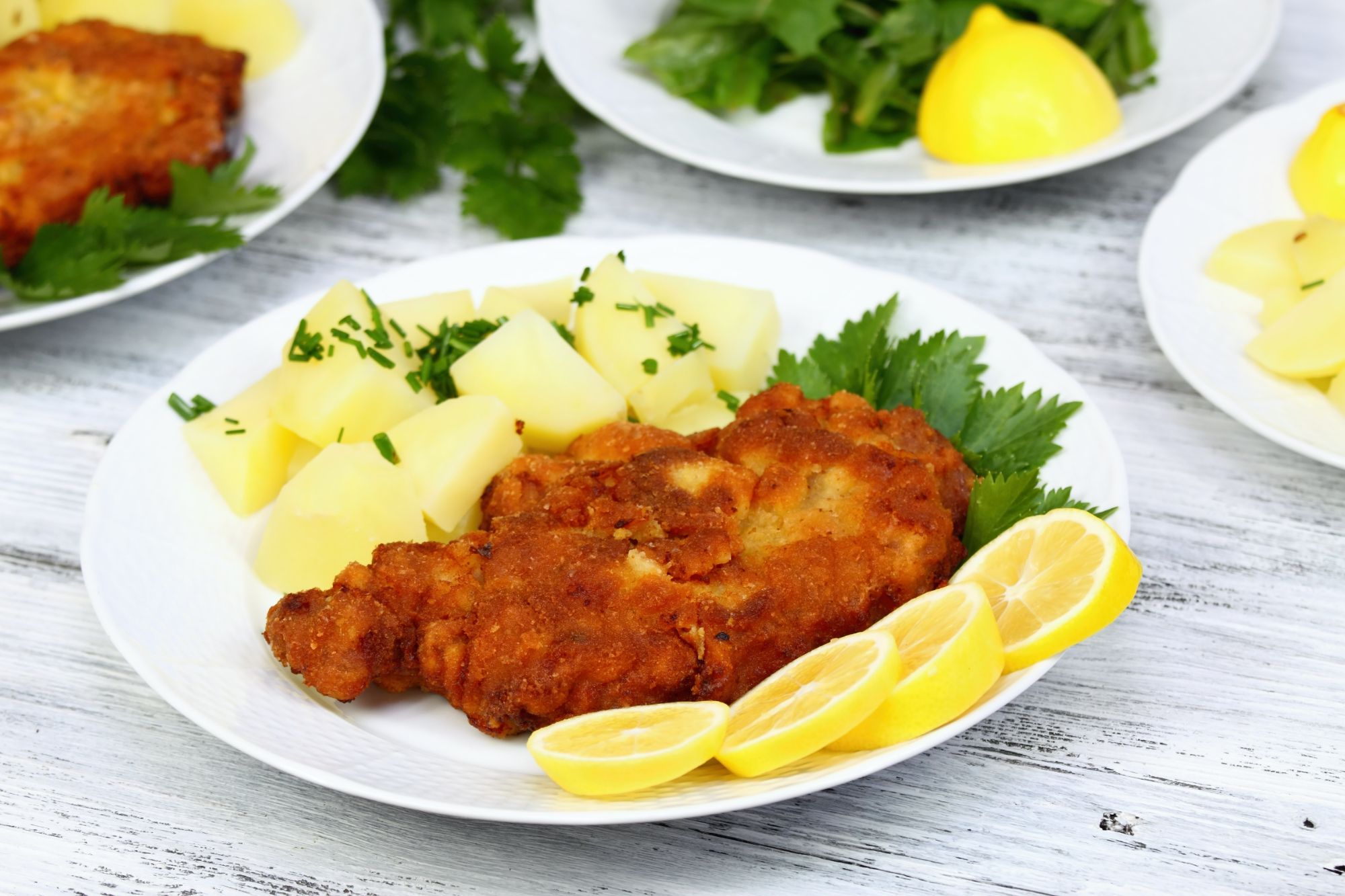Złocisty kotlet schabowy na talerzu z ziemniakami, cytryną i świeżą kolendrą.