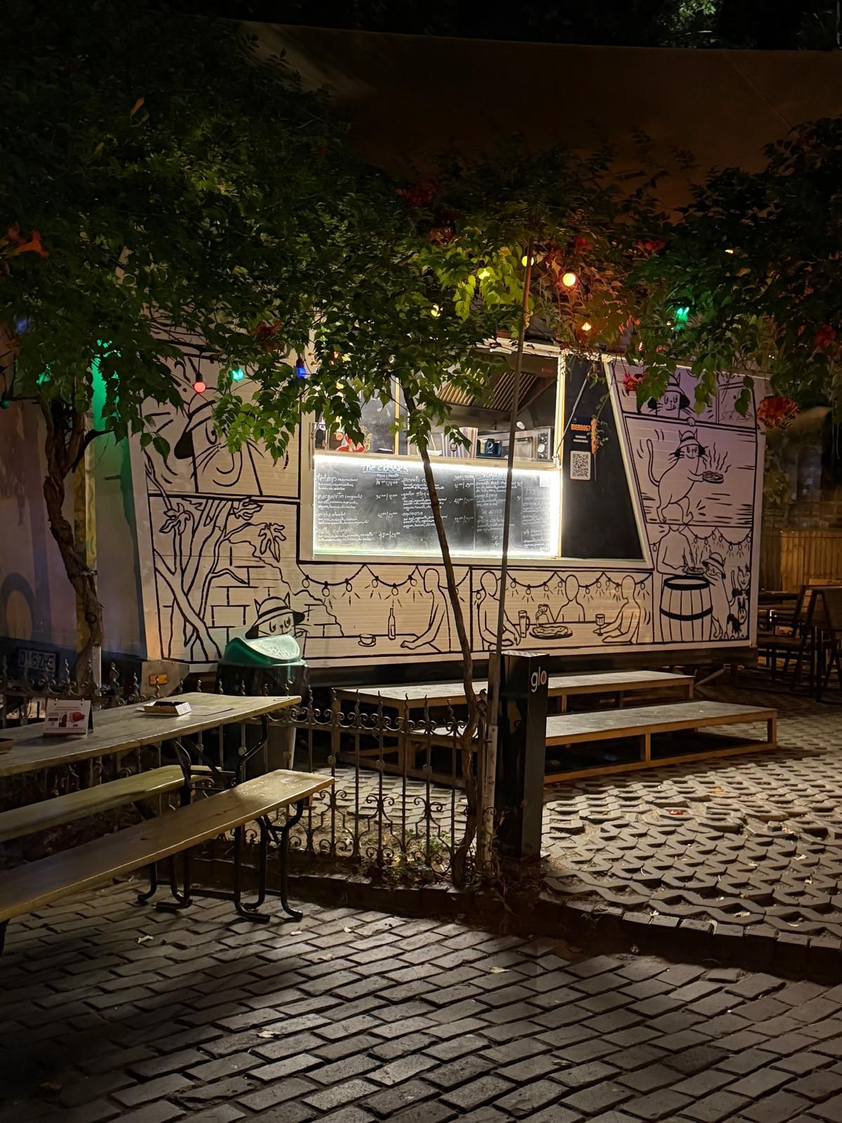 Bară exterioară iluminată cu LED-uri, cu un camion de mâncare decorat artistic și mese de lemn în jur.