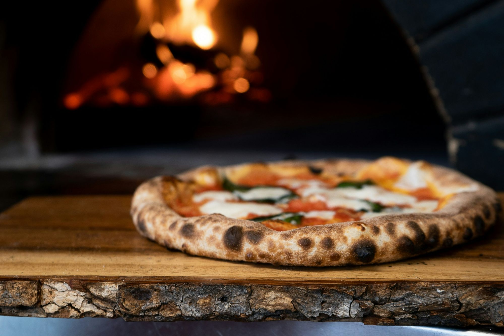 Pyszna pizza margherita na drewnianej desce, w tle żar pieca opalanego drewnem.