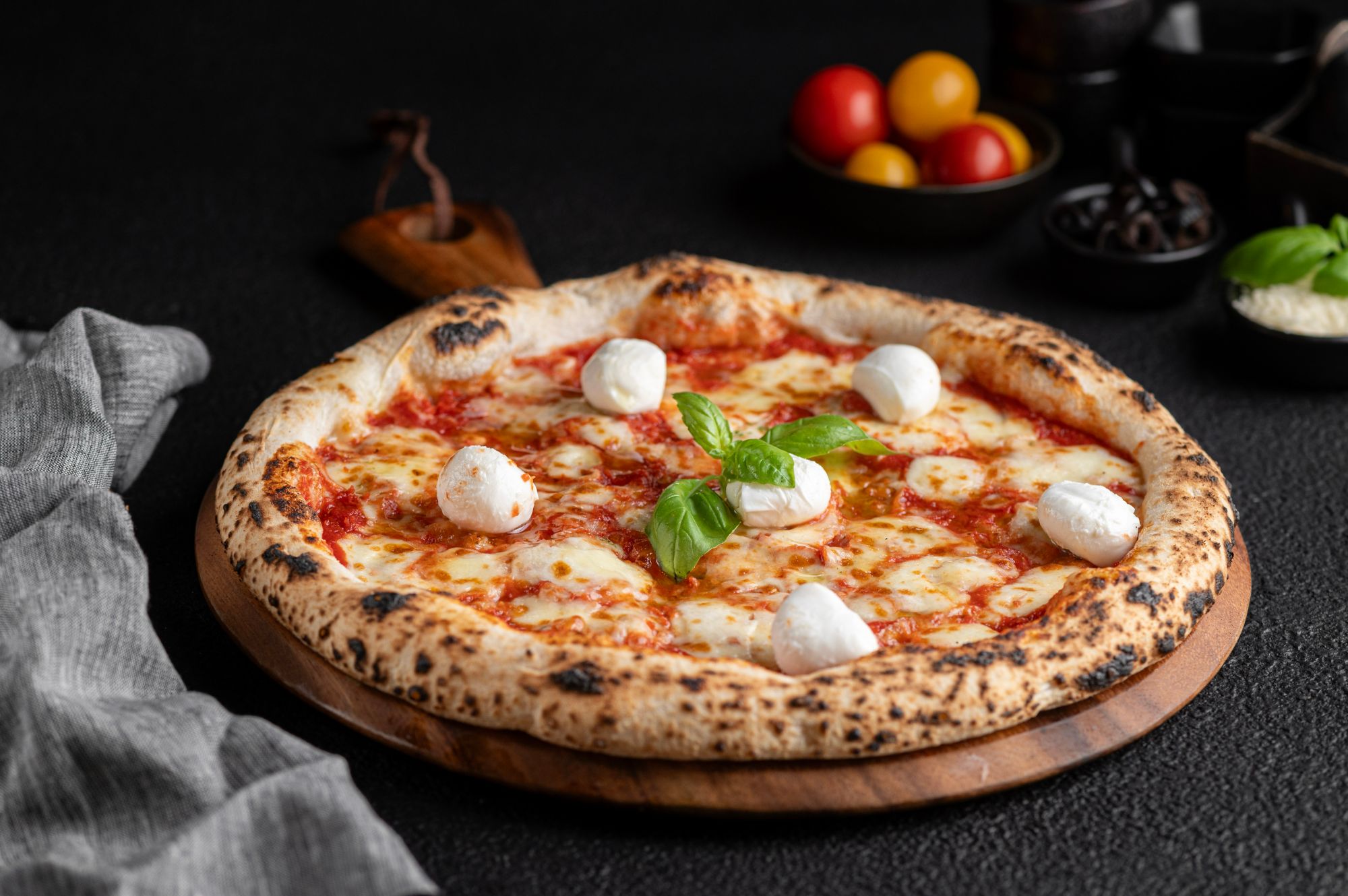 Pizza margherita z serem, bazylią i mini kulkami mozzarelli na drewnianej desce, z kolorowymi pomidorami w tle.