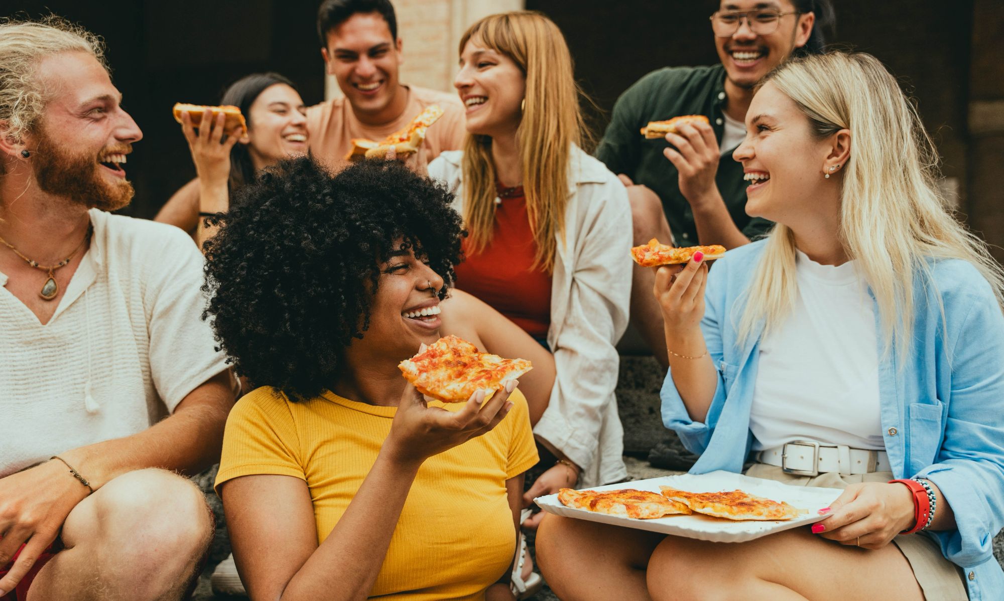 Grupa przyjaciół wesoło dzieli się pizzą na schodach, uśmiechając się i rozmawiając.