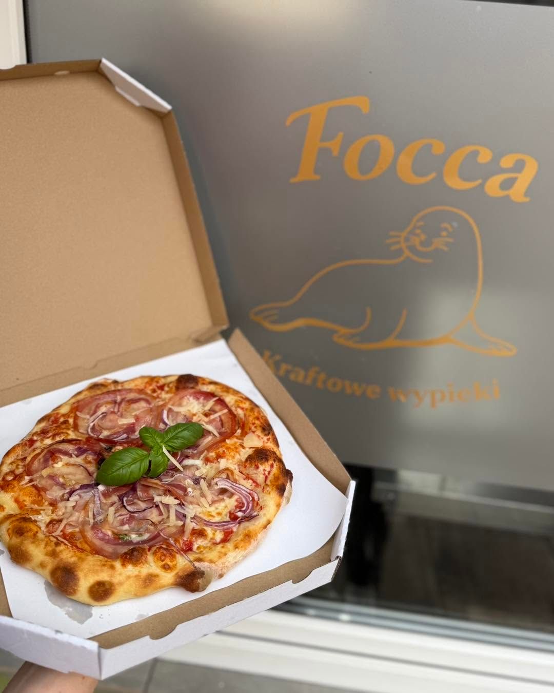 Pizza w opakowaniu, z plasterkami szynki i bazylią, przed napisem "Focca Kraftowe Wypieki".