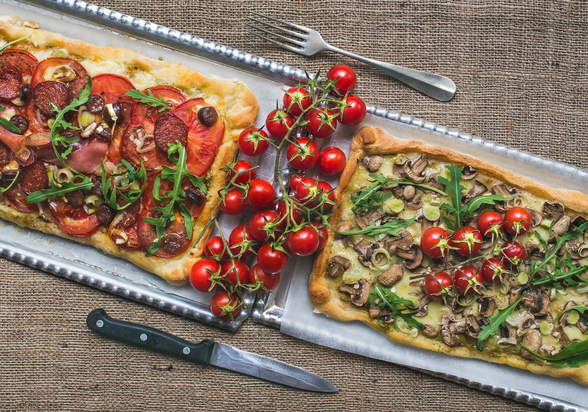 Dwie pizze na blaszce: jedna z pepperoni i pomidorami, druga z pieczarkami i rukolą, obok pomidory koktajlowe.