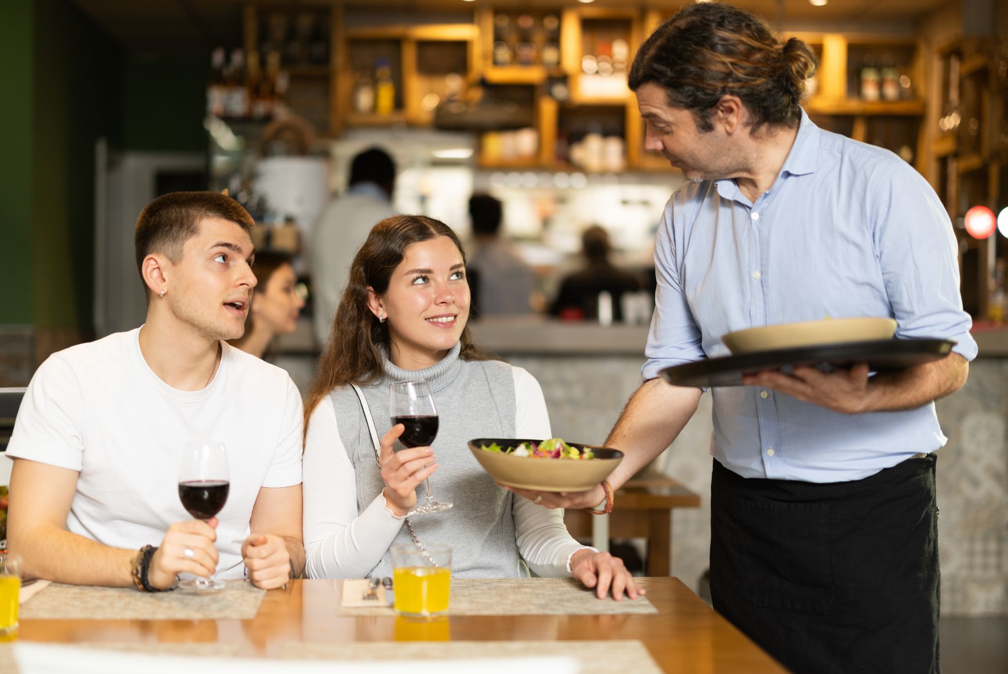 Kelner podaje danie parze w restauracji, oboje mają lampki wina, a w tle widać innych gości.