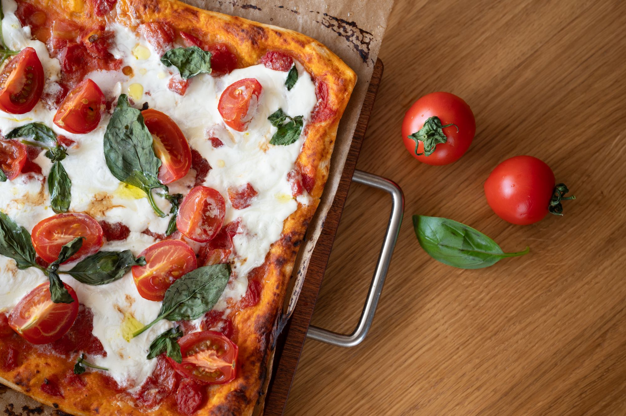 Pizza z sosem pomidorowym, serem mozzarella, pomidorami i bazylią na drewnianym blacie, obok kilka całych pomidorów.