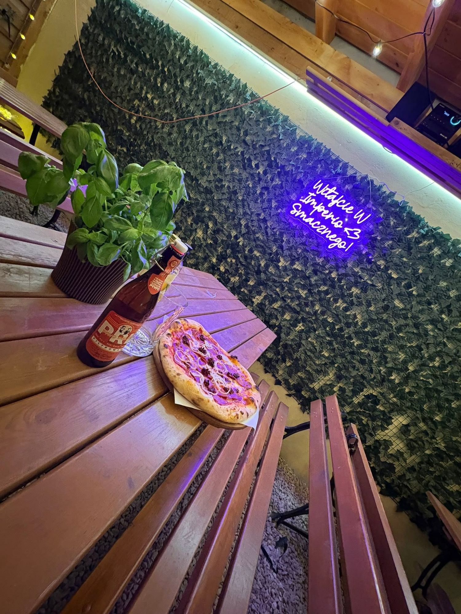 Pizza na stole z piwem w przytulnej restauracji z roślinnym tłem i neonowym napisem.
