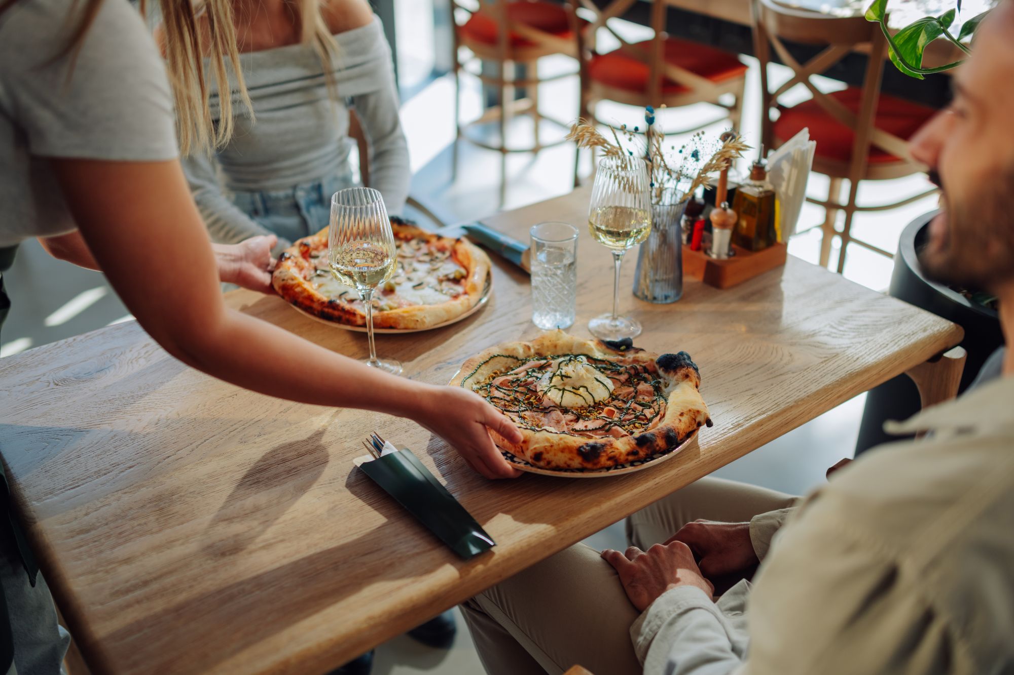 Kobieta serwuje pizze przy stole w restauracji, z winem i dekoracjami w tle.