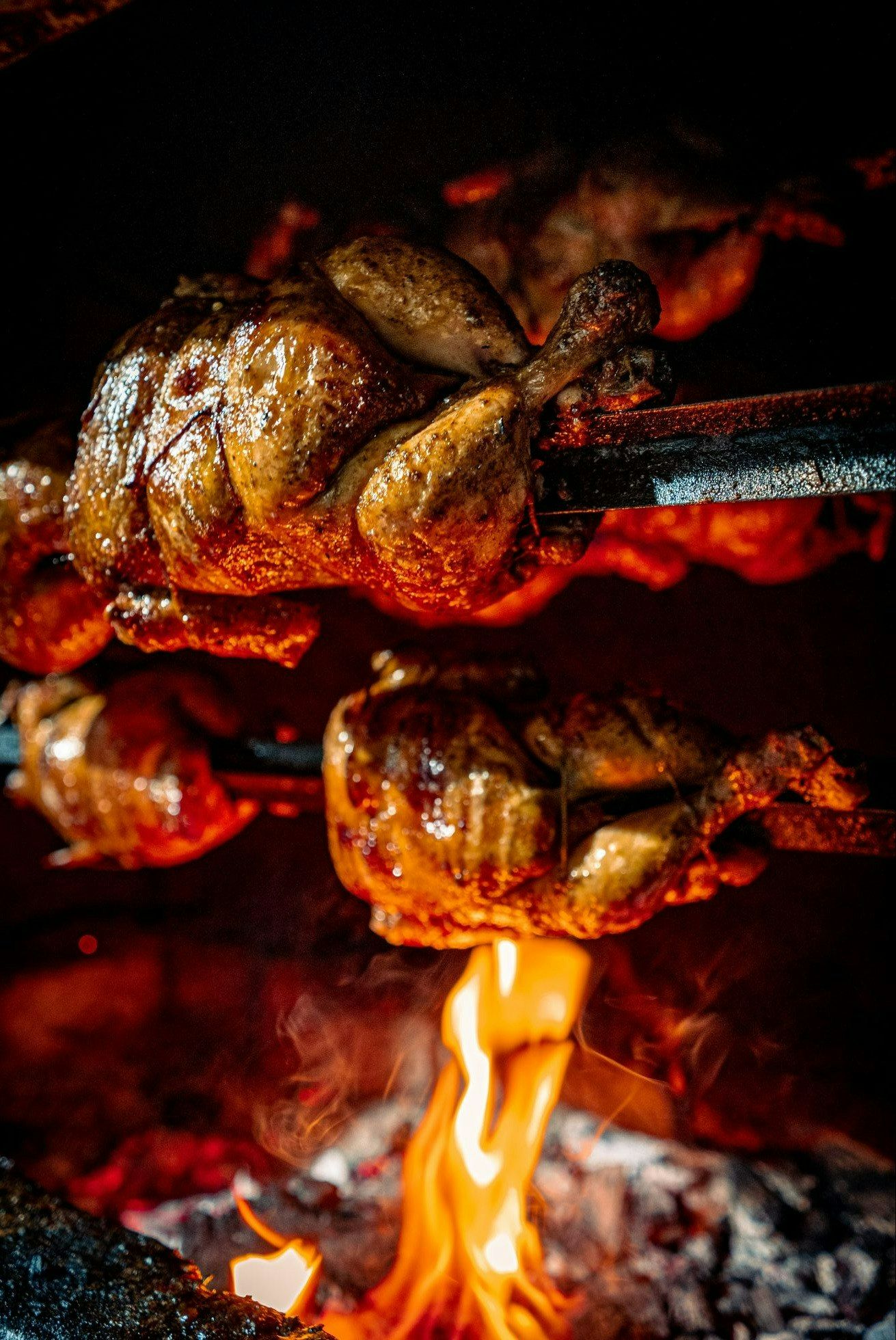 Pui la rotisor, rumenit, gătit pe un foc viu, cu flăcări și cărbuni în fundal.
