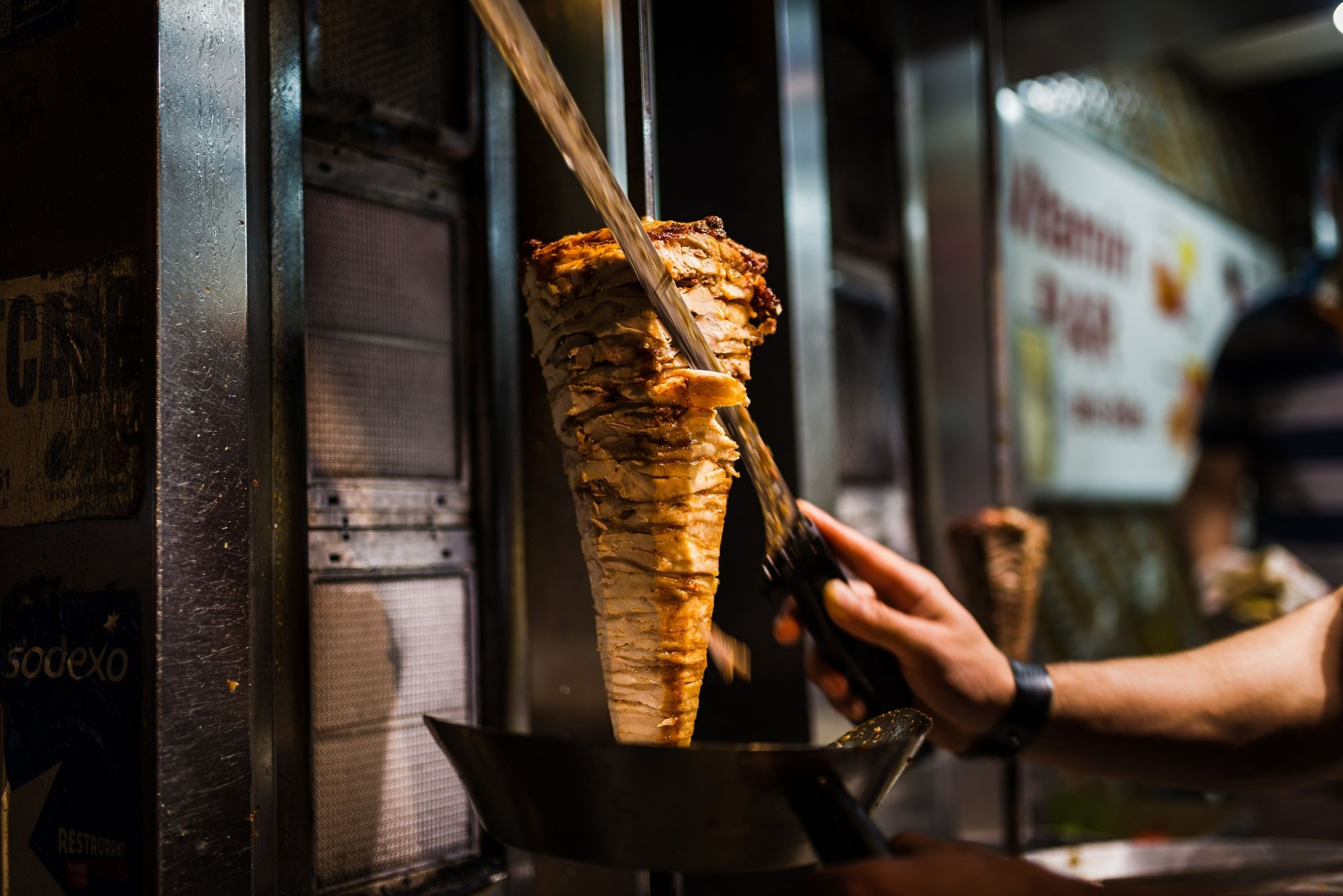 O persoană taie carne dintr-un con de kebab la un stand de street food, cu un fundal animat.