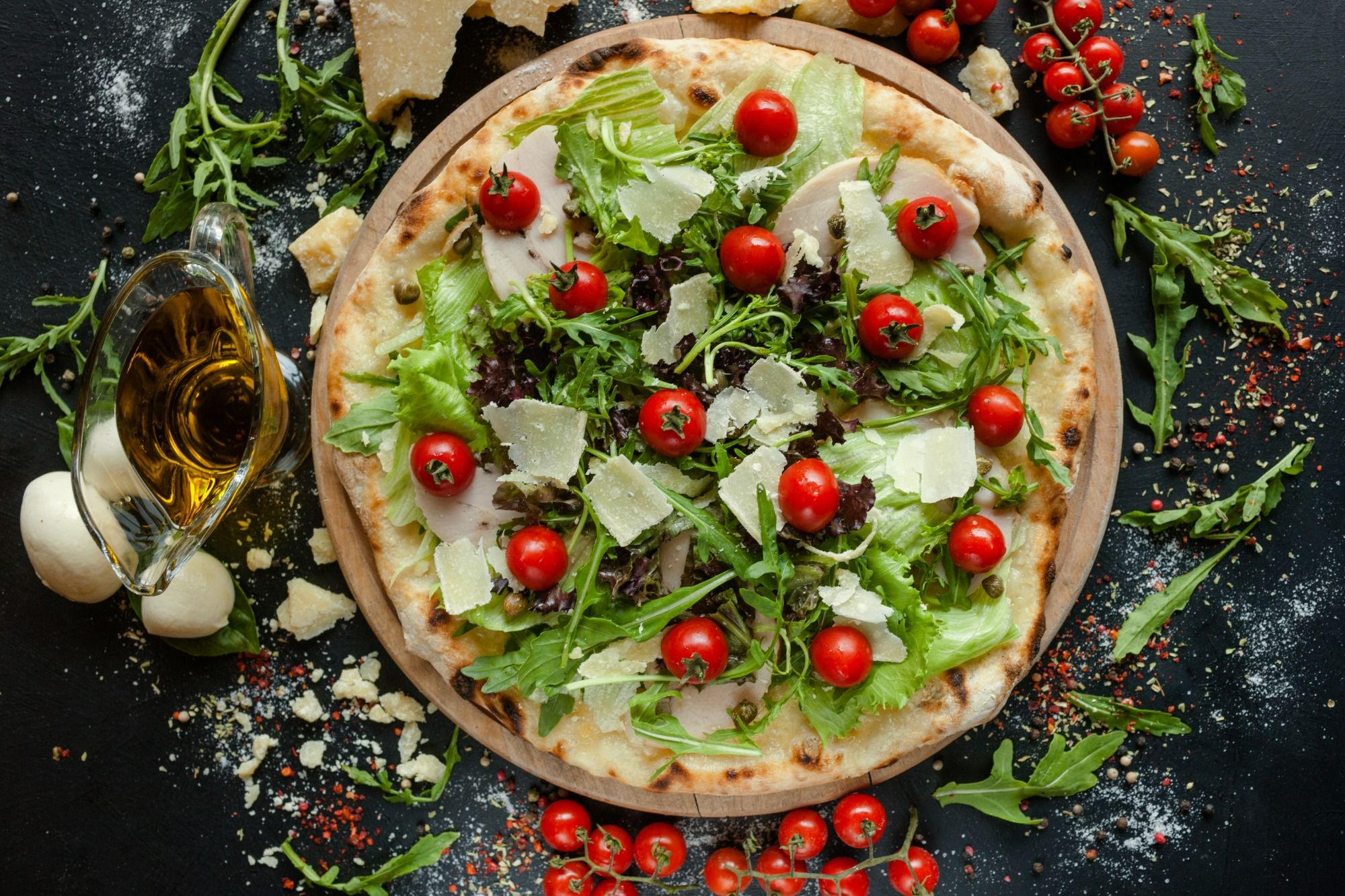 Pizza z sałatą, pomidorkami koktajlowymi i serem, otoczona oliwą i przyprawami na czarnym tle.