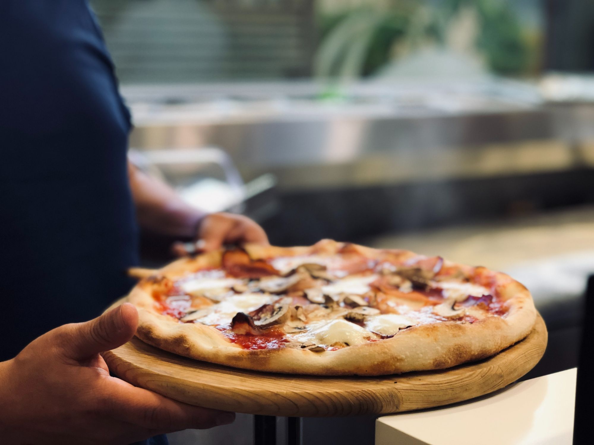 Osoba trzymająca pizzę na drewnianym talerzu z serem, pieczarkami i szynką w restauracji.