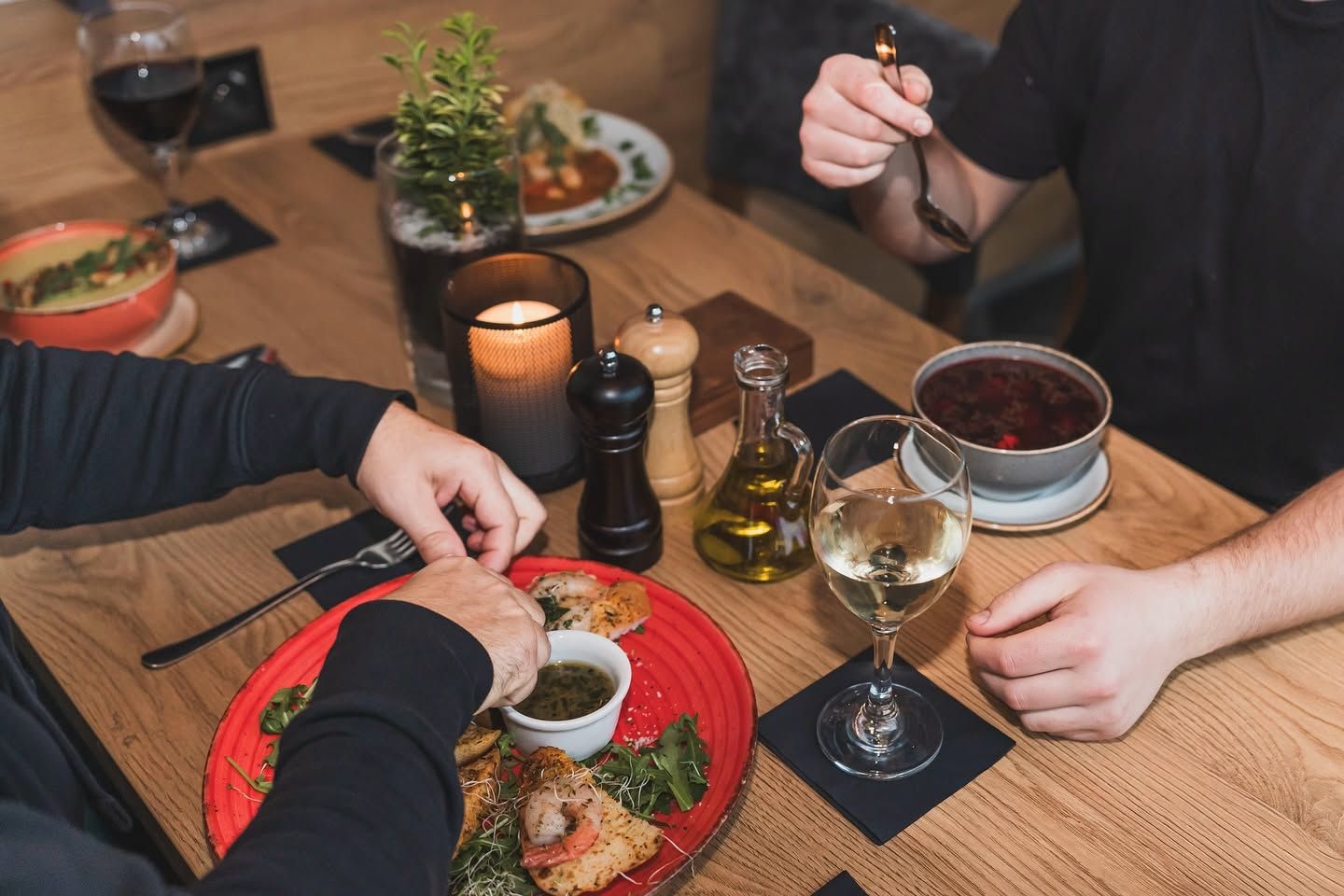 Osoby jedzące posiłek w restauracji, z talerzem przystawek, winem i świecą na stole.