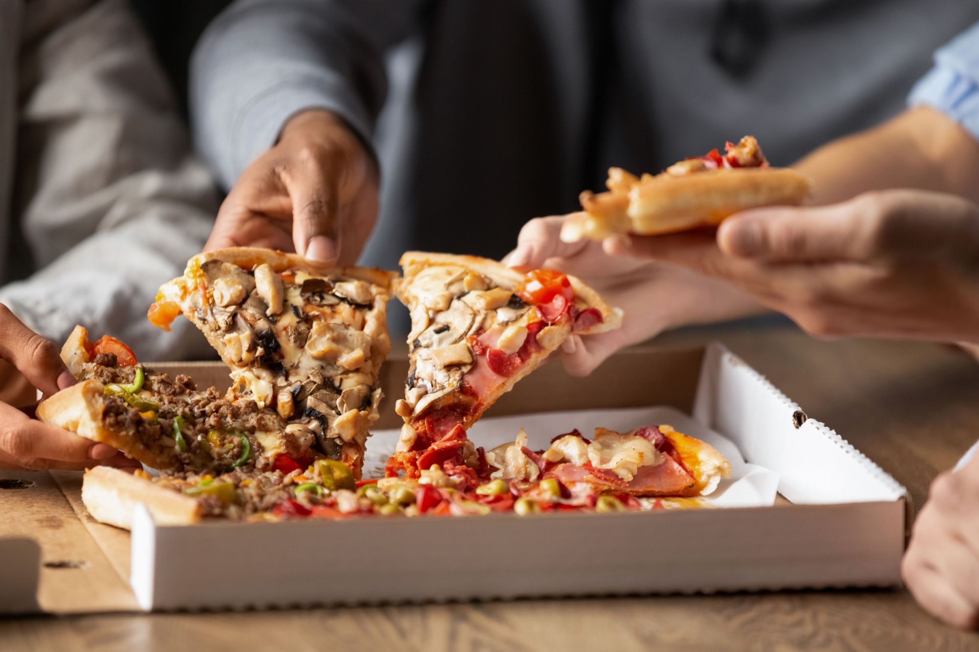 Grupa osób dzieli się kawałkami pizzy w kartonowym opakowaniu na drewnianym stole.