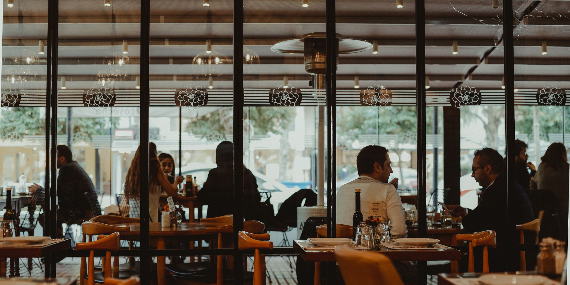 Wnętrze restauracji z klientami siedzącymi przy stolikach, przeszklona ściana i nowoczesny wystrój.
